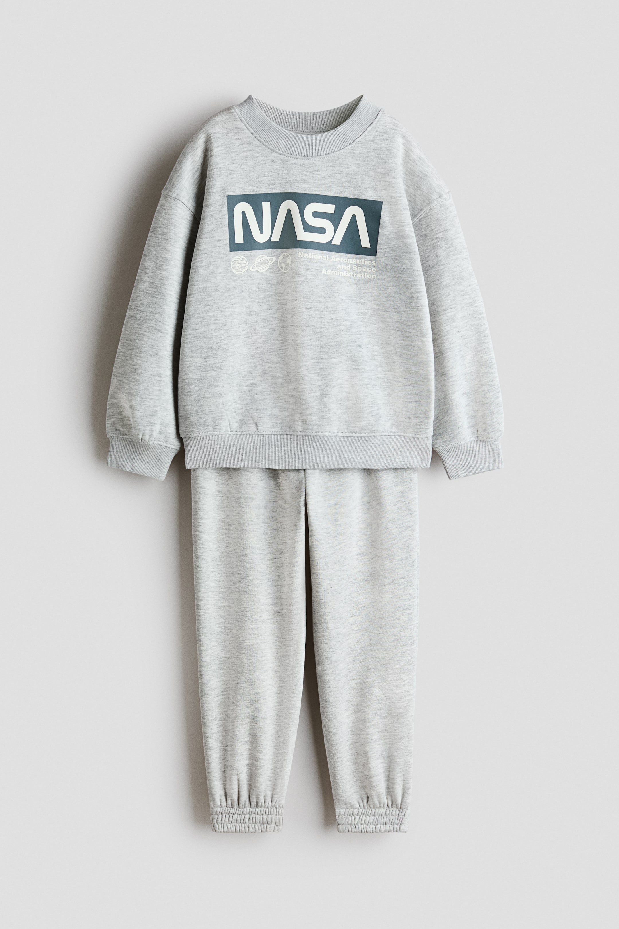 Grotere afbeelding bekijken: Een lichtgrijs gemêleerd trainingspak bestaande uit een sweatshirt met ronde hals en bijpassende joggingbroek, beide voorzien van geribde boorden en zomen. Het sweatshirt toont een grote rechthoekige afbeelding op de borst, met daarop 'NASA' in wit op een donker teal achtergrond, naast kleinere witte planetaire contouren en de volledige tekst 'National Aeronautics and Space Administration'.