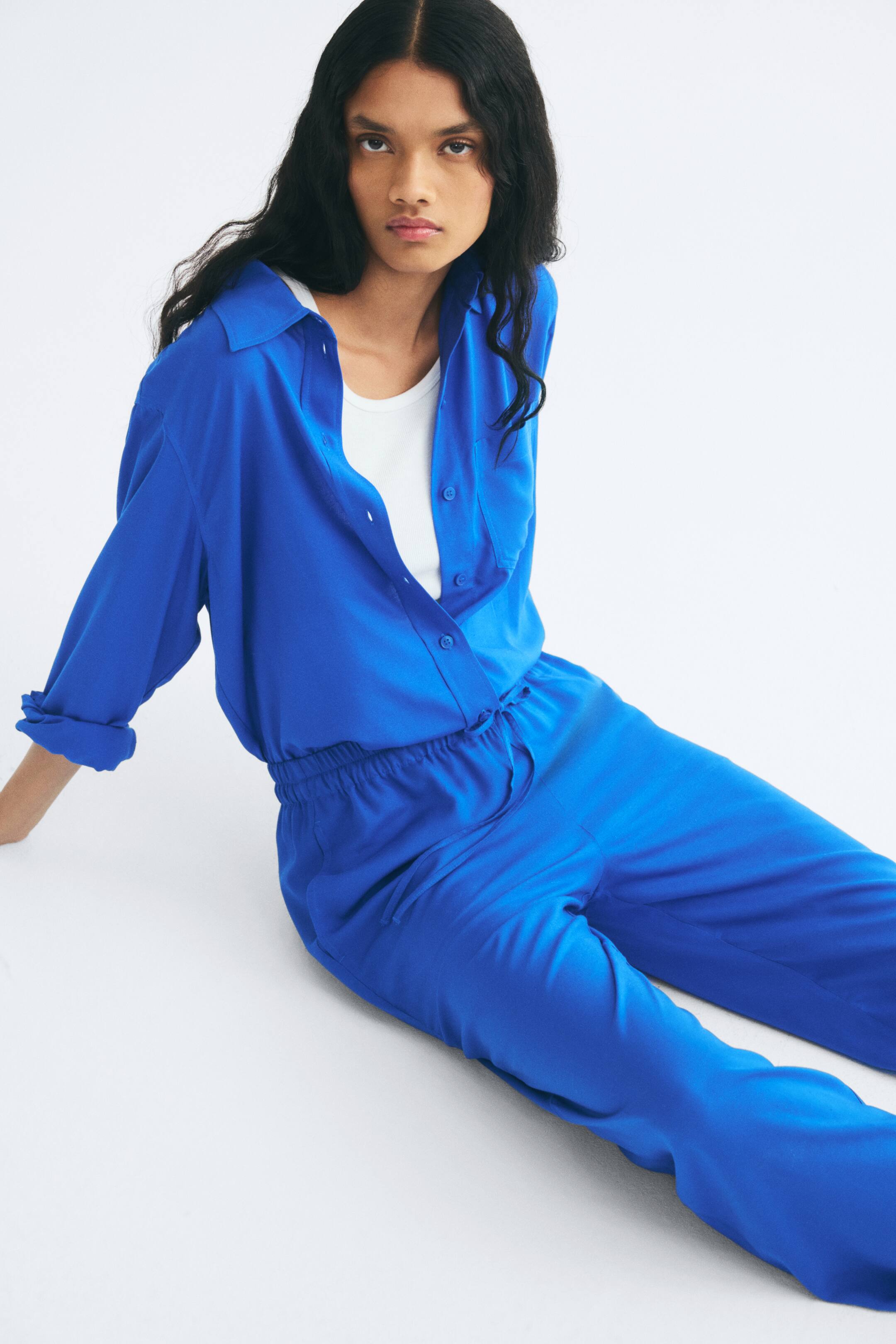 View larger image: Bright blue wide-leg pants featuring an elasticated drawstring waist are worn by the model while seated. The pants are paired with a matching blue long-sleeved button-up shirt, worn open over a white tank top.