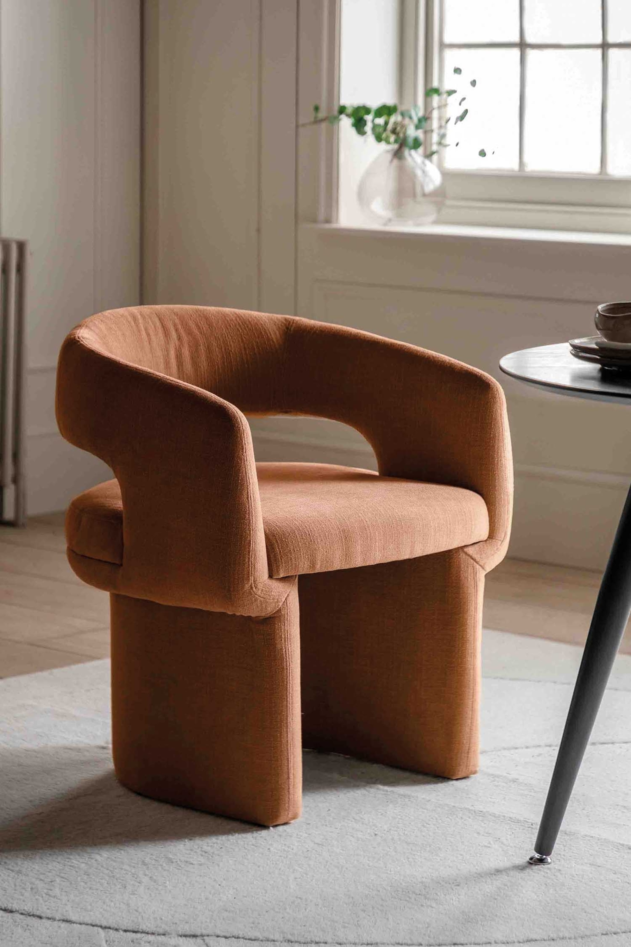 View larger image: A terracotta-toned upholstered armchair is showcasing its front-left angle, featuring a continuous curved backrest and arms with a central cut-out, and a solid upholstered block-like base.
