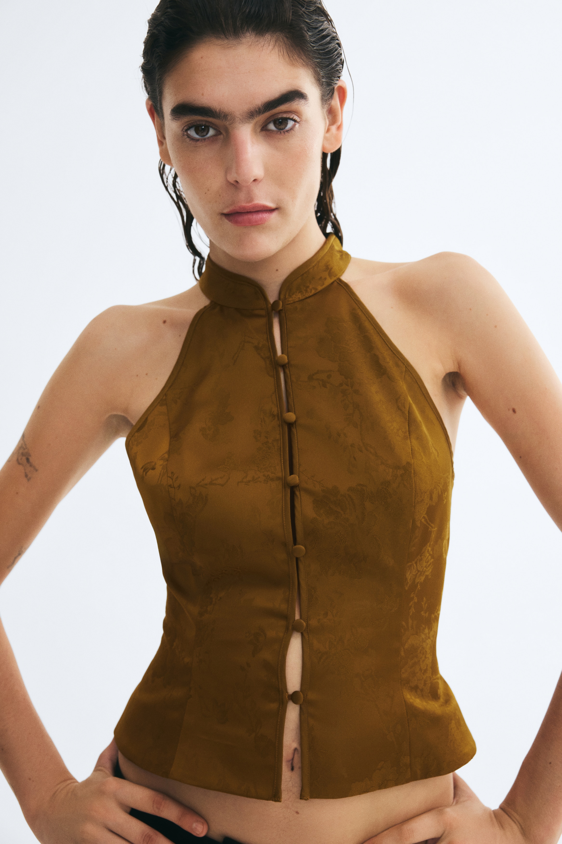 A mustard-brown halterneck top is facing front, showcasing a subtle floral jacquard pattern and featuring a row of small, fabric-covered buttons down the centre. The sleeveless top is displaying a high, structured collar and tailored princess seams that extend to the straight hem.