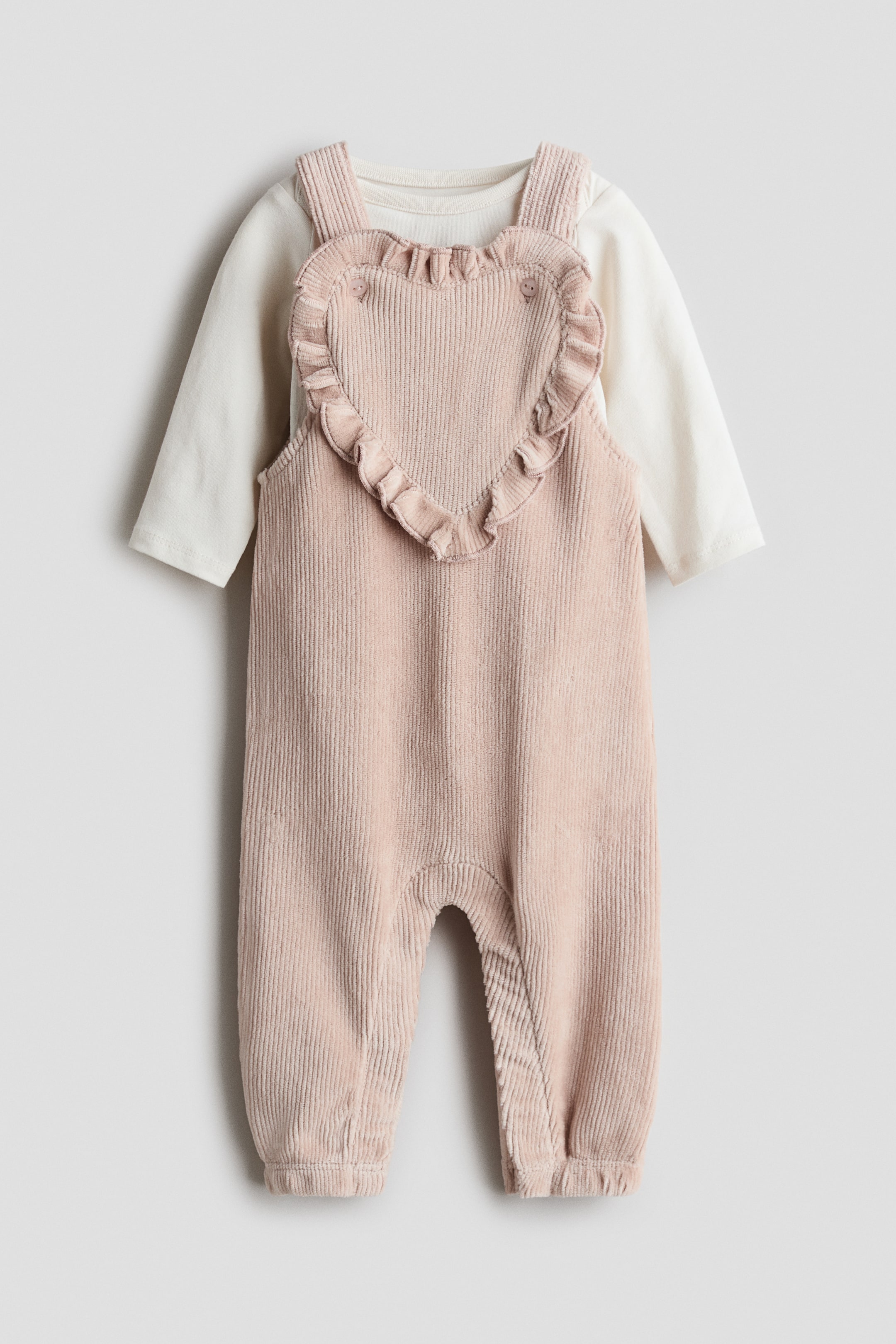View larger image: A baby's two-piece outfit is presenting FRONT, featuring a pale pink corduroy overall with a heart-shaped bib and an off-white long-sleeved top layered underneath. The overall is showcasing wide straps, ruffles outlining the heart-shaped bib, and elasticated ankle cuffs.