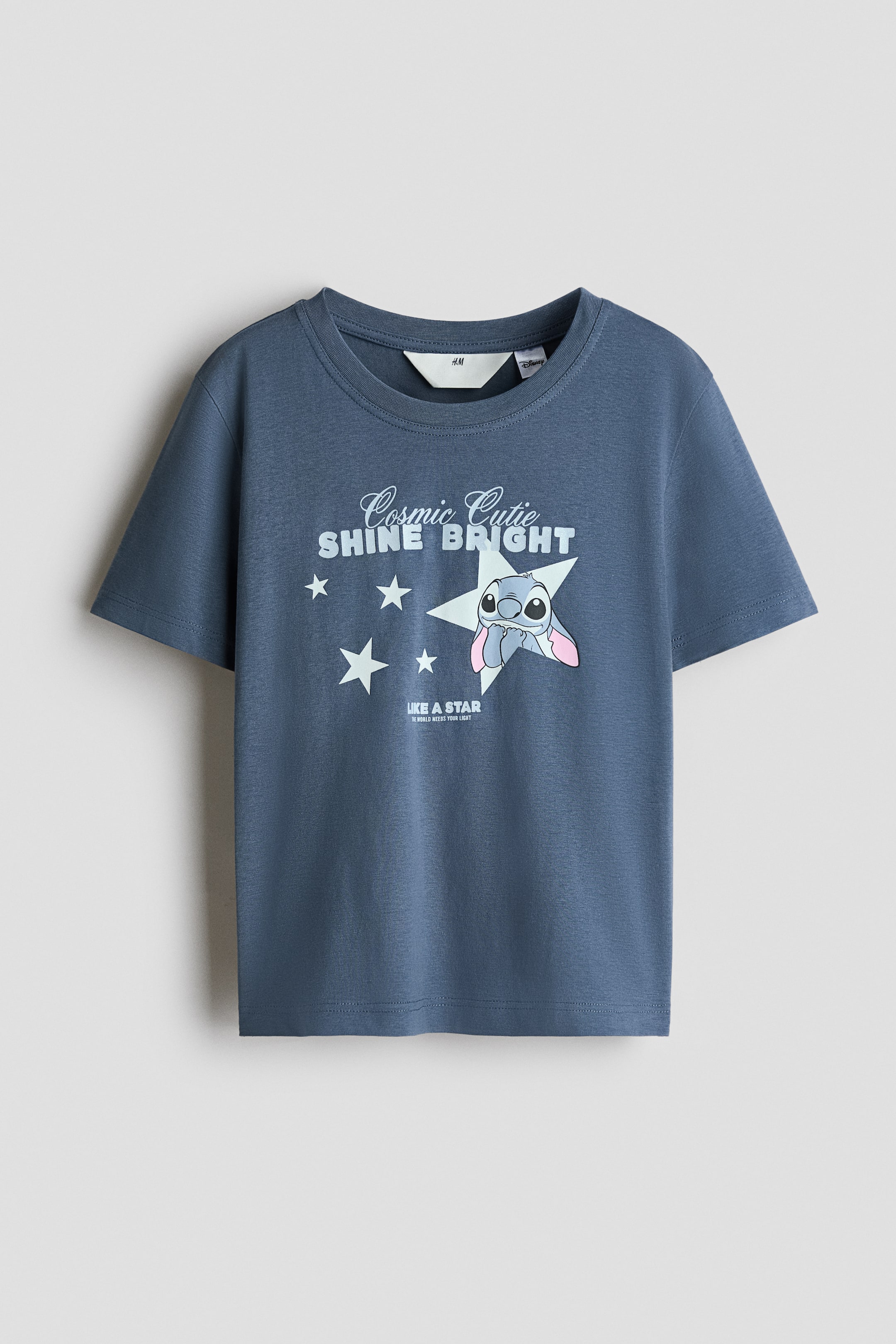 View larger image: A slate blue, short-sleeved crew neck t-shirt featuring a front-facing graphic of Stitch peeking from behind a white star. The graphic includes the text "Cosmic Cutie SHINE BRIGHT" and "LIKE A STAR," along with smaller white stars.