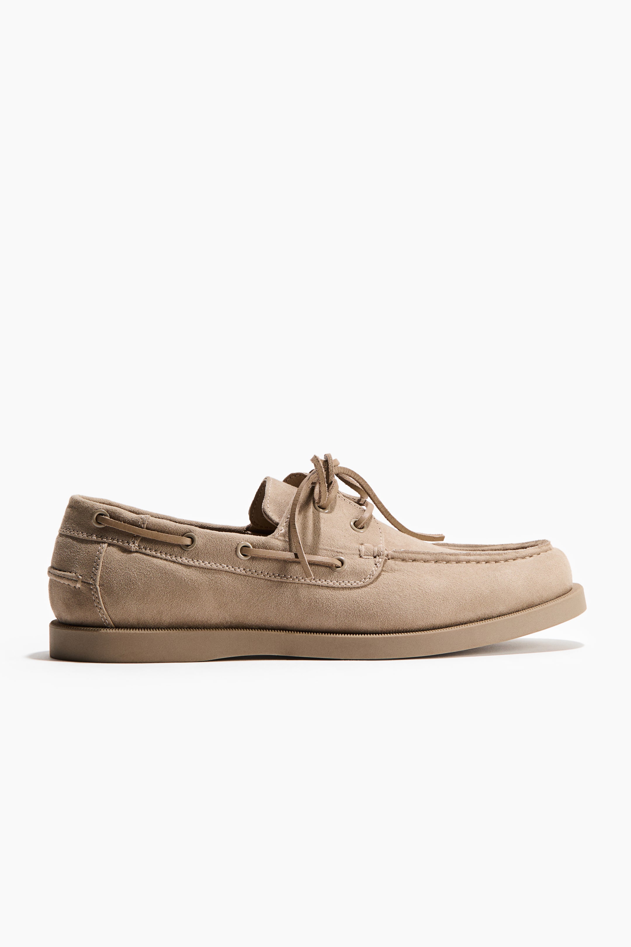 Agrandir l'image: Une chaussure bateau en daim beige est présentée de profil, avec un bout de style mocassin. Des lacets en cuir sont enfilés à travers des œillets métalliques sur le dessus et s'étendent autour du col, la chaussure reposant sur une semelle plate assortie de couleur beige.