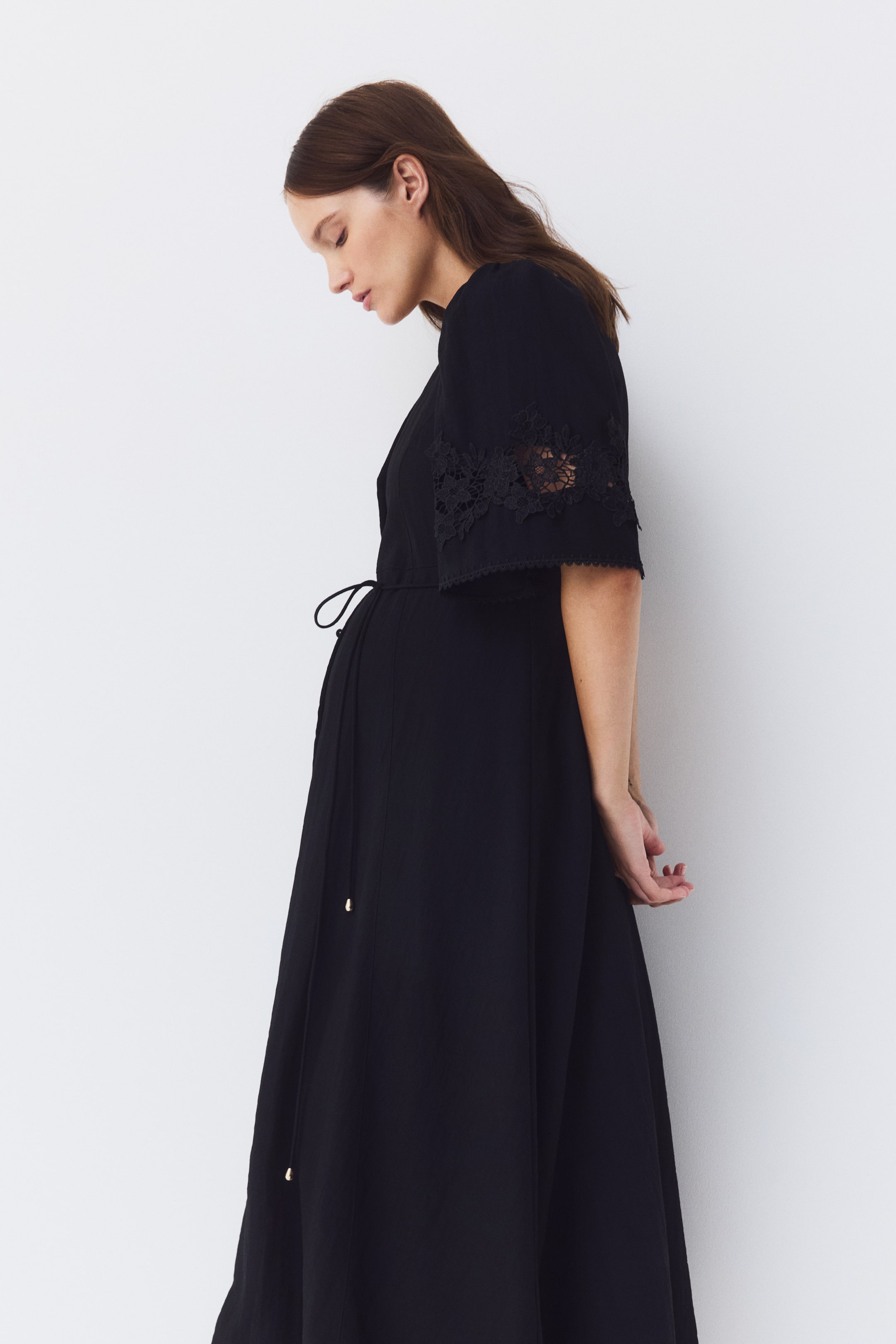 Ver imagen más grande: La modelo lleva un vestido largo negro. Está diseñado con un escote en V, mangas abullonadas hasta el codo detalladas con broderie anglaise y una falda que se ensancha suavemente, mientras que un cinturón de lazo con extremos de cuentas ajusta la cintura.