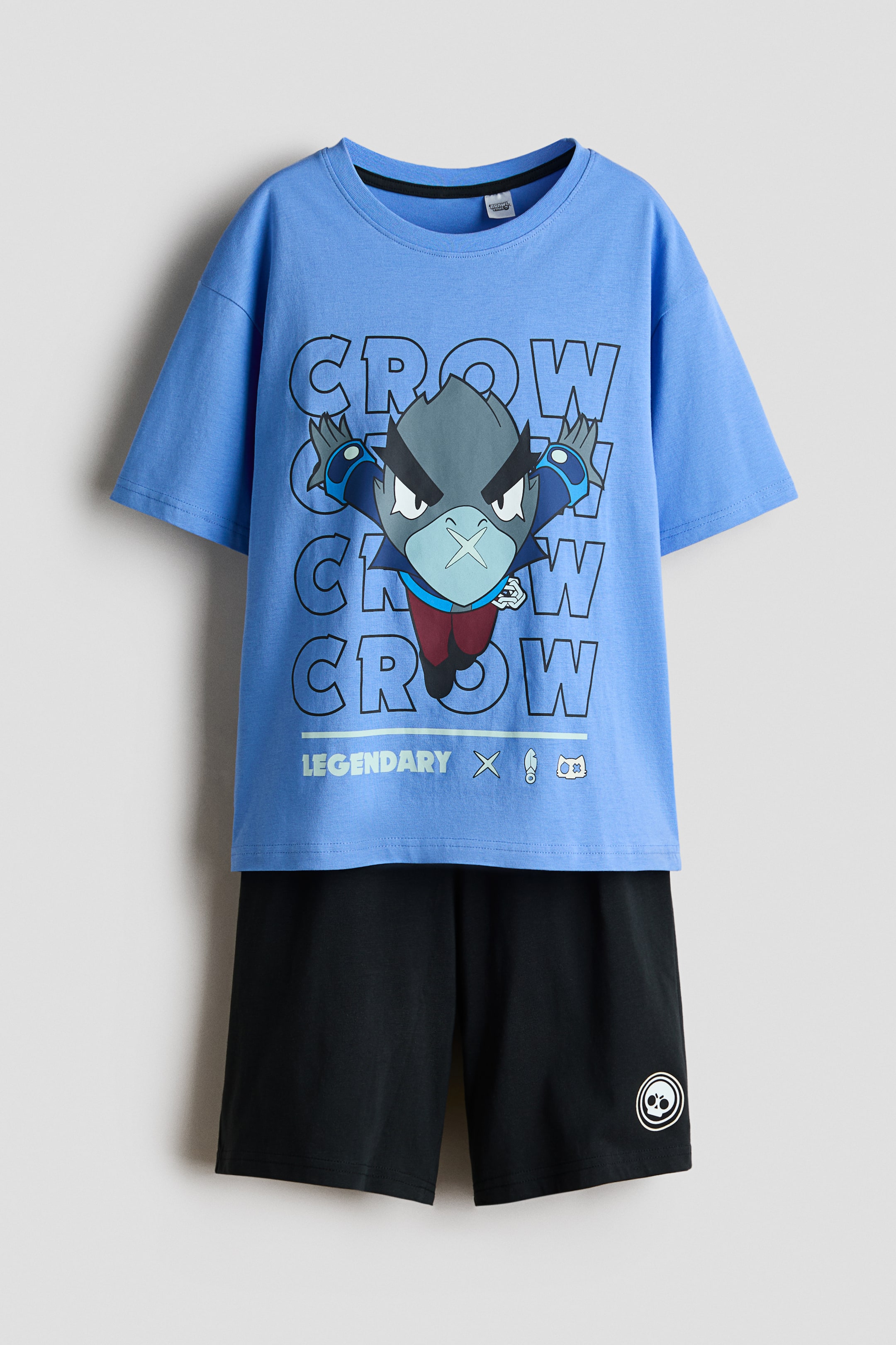 View larger image: Presenting a pyjama set, comprising a T-shirt and shorts crafted from soft cotton jersey, shown in a front view. The blue T-shirt is adorned with a character print motif featuring the text "CROW," complemented by a round, ribbed neckline and subtly dropped shoulders. The black shorts are designed with an elasticated waist and showcase a white skull logo on the lower left leg.