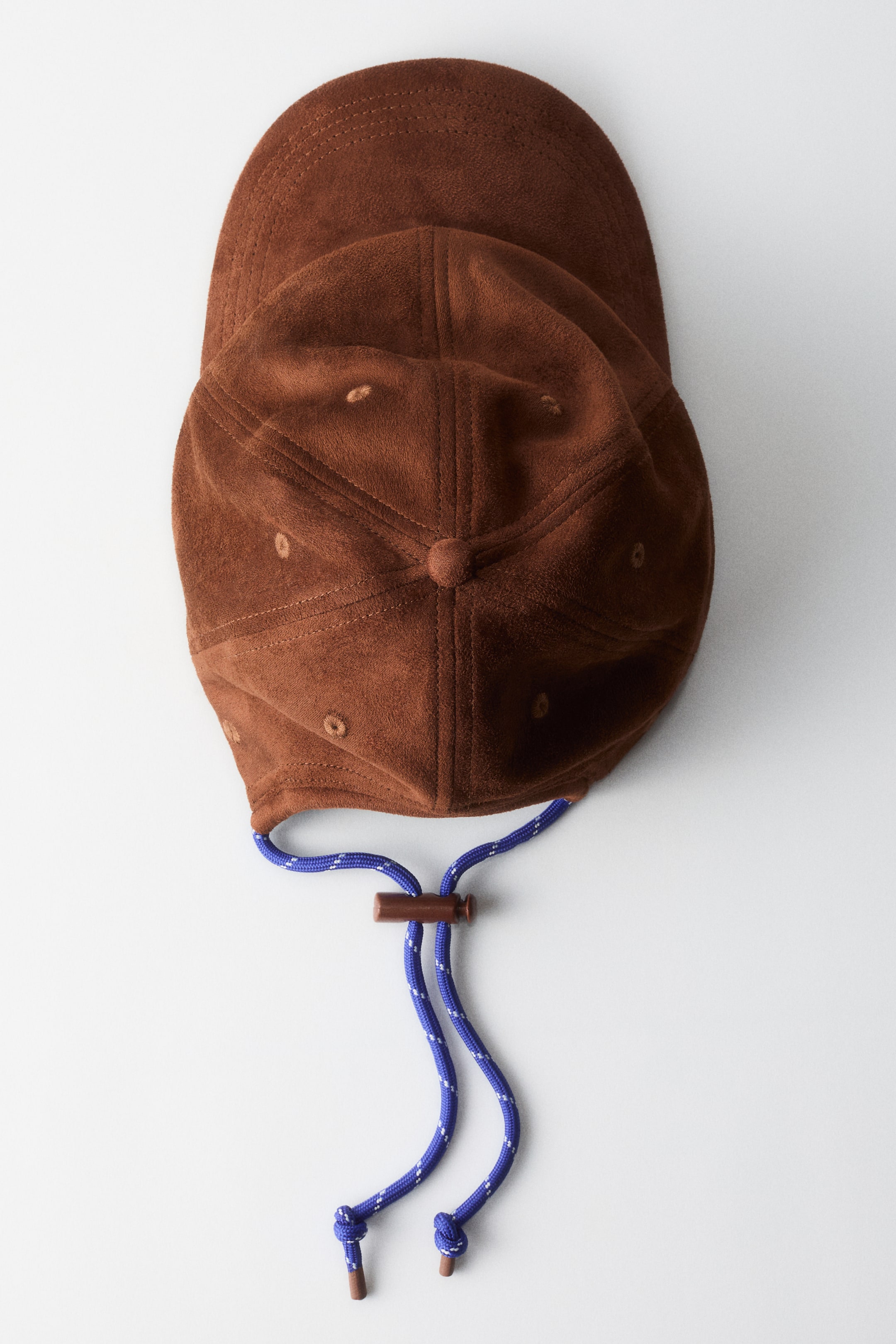 Ampliar la imagen: Una gorra de color marrón con apariencia de gamuza se encuentra plana, vista desde arriba, con una visera curva y una corona de seis paneles con seis ojales bordados y un botón superior. Un cordón azul y blanco moteado con un tope de madera y cuentas de madera en los extremos se extiende desde la parte trasera de la gorra.