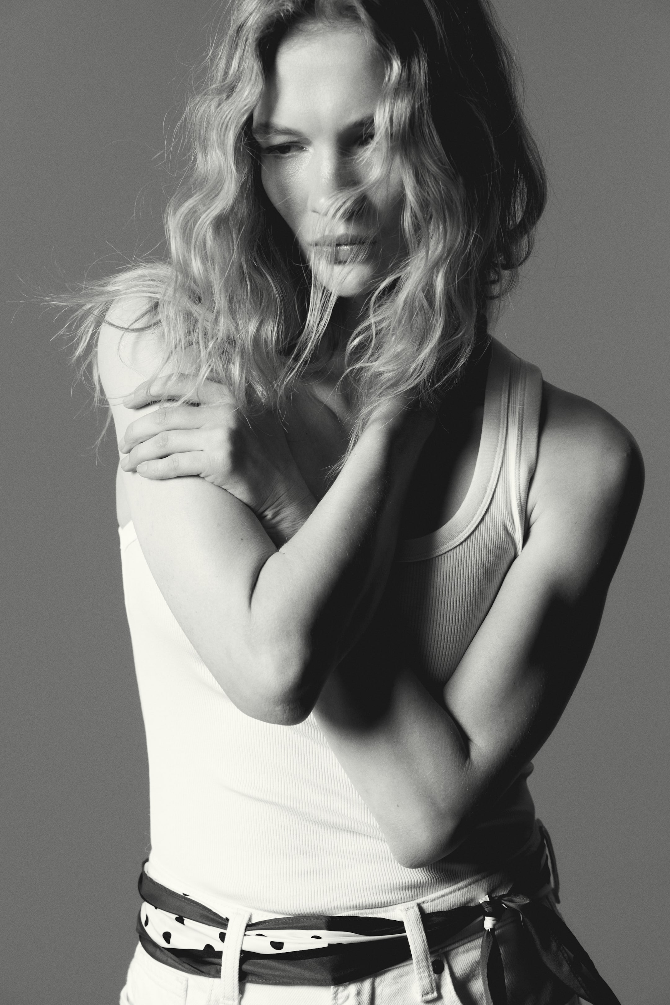 Nagyobb kép megtekintése: Egy fehér bordázott trikó szorosan illeszkedik a testhez, kerek nyakkivágással és széles vállpántokkal rendelkezik. Fehér nadrággal van viselve, és egy fekete-fehér pöttyös sálat használnak övként a derék körül.