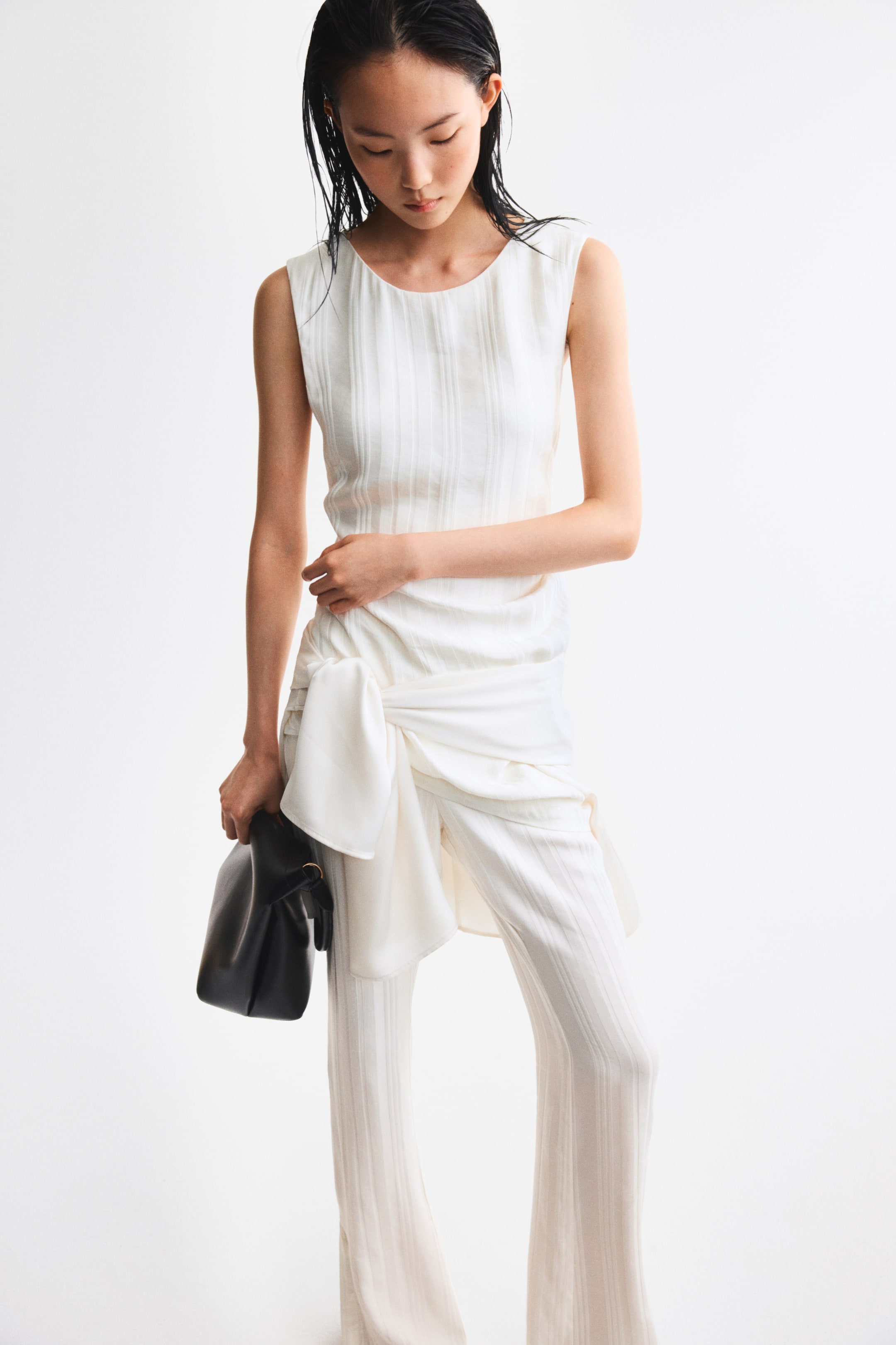 View larger image: A long, sleeveless top with a textured weave and a round neckline is worn by the model. It features pleats at one side of the waist, creating an asymmetric hem, and the model is touching this draped area.