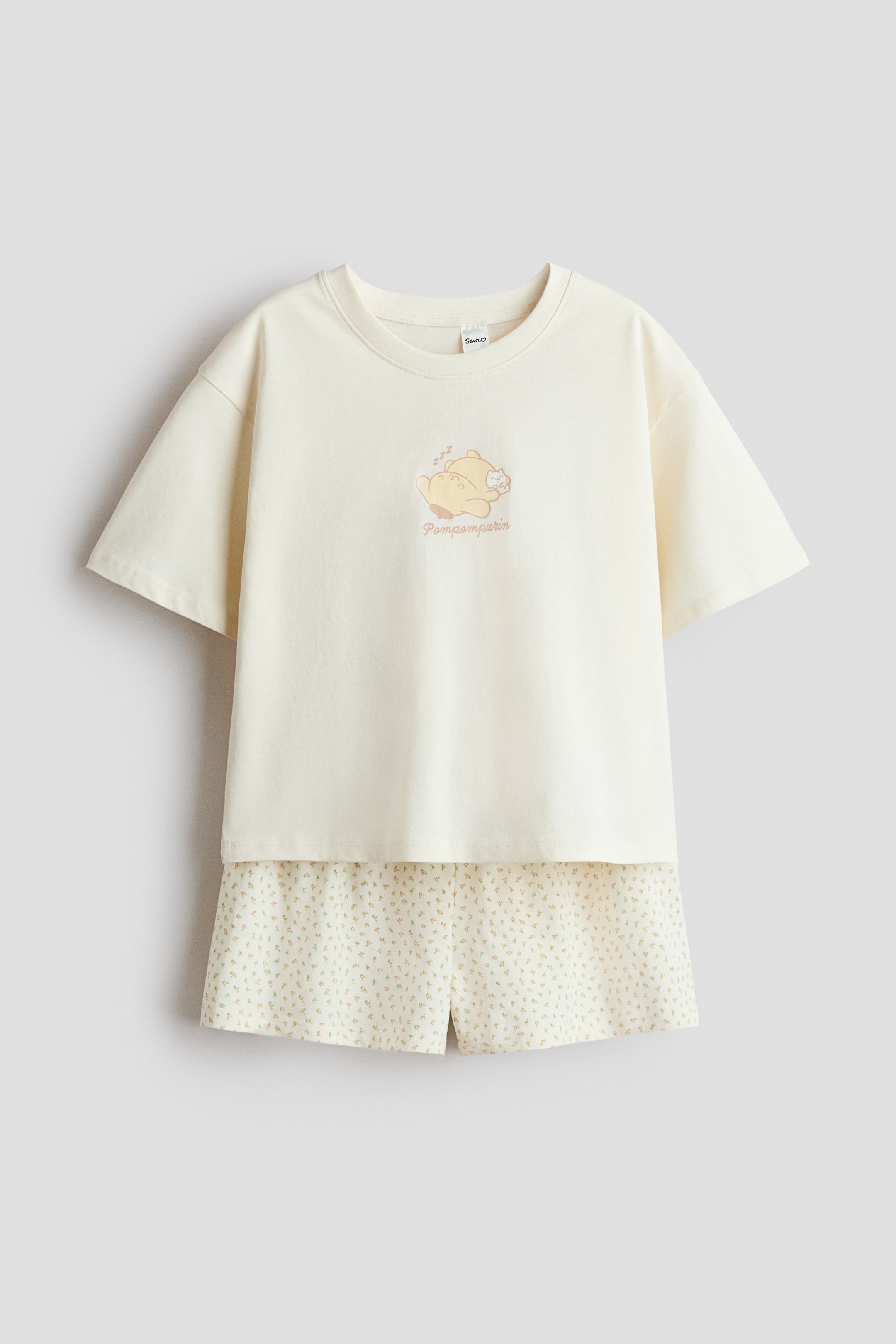 Größeres Bild ansehen: Ein Paar Pyjamas aus weichem, bedrucktem Baumwolljersey wird von vorne präsentiert und besteht aus einem cremefarbenen T-Shirt und passenden Shorts. Das T-Shirt verfügt über einen gerippten Halsausschnitt, überschnittene Schultern und einen schlafenden Pompompurin-Charakter mit Text auf der Vorderseite, während die Shorts einen elastischen Bund und einen durchgehenden kleinen Blumenprint aufweisen.