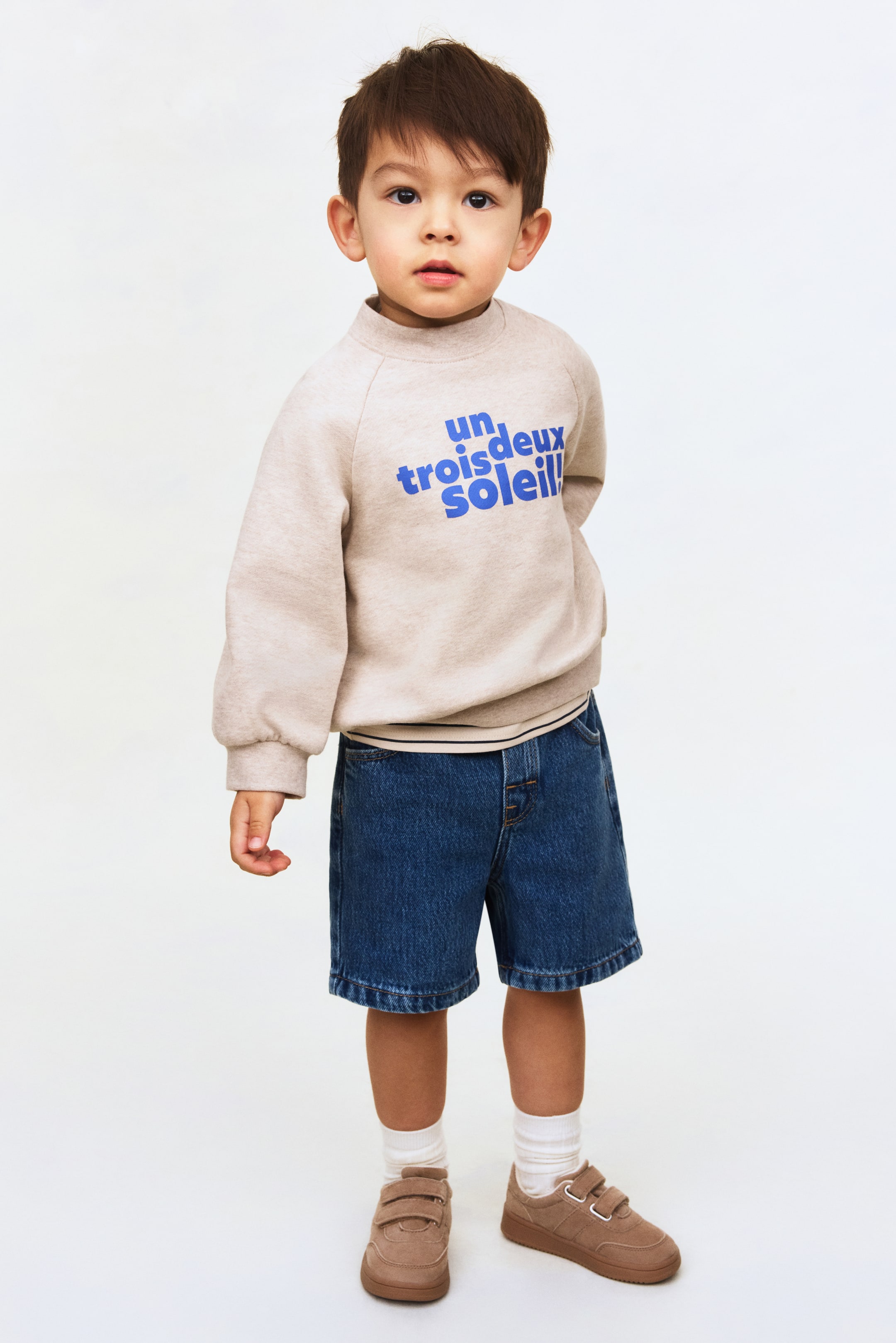 Prikaži veću sliku: Bež dukserica sa polurolkom i plavim natpisom "un trois deux soleil!" na prednjem delu. Ima raglan rukave sa rebrastim manžetnama i rebrastim donjim rubom i nosi se preko tanke bele majice.