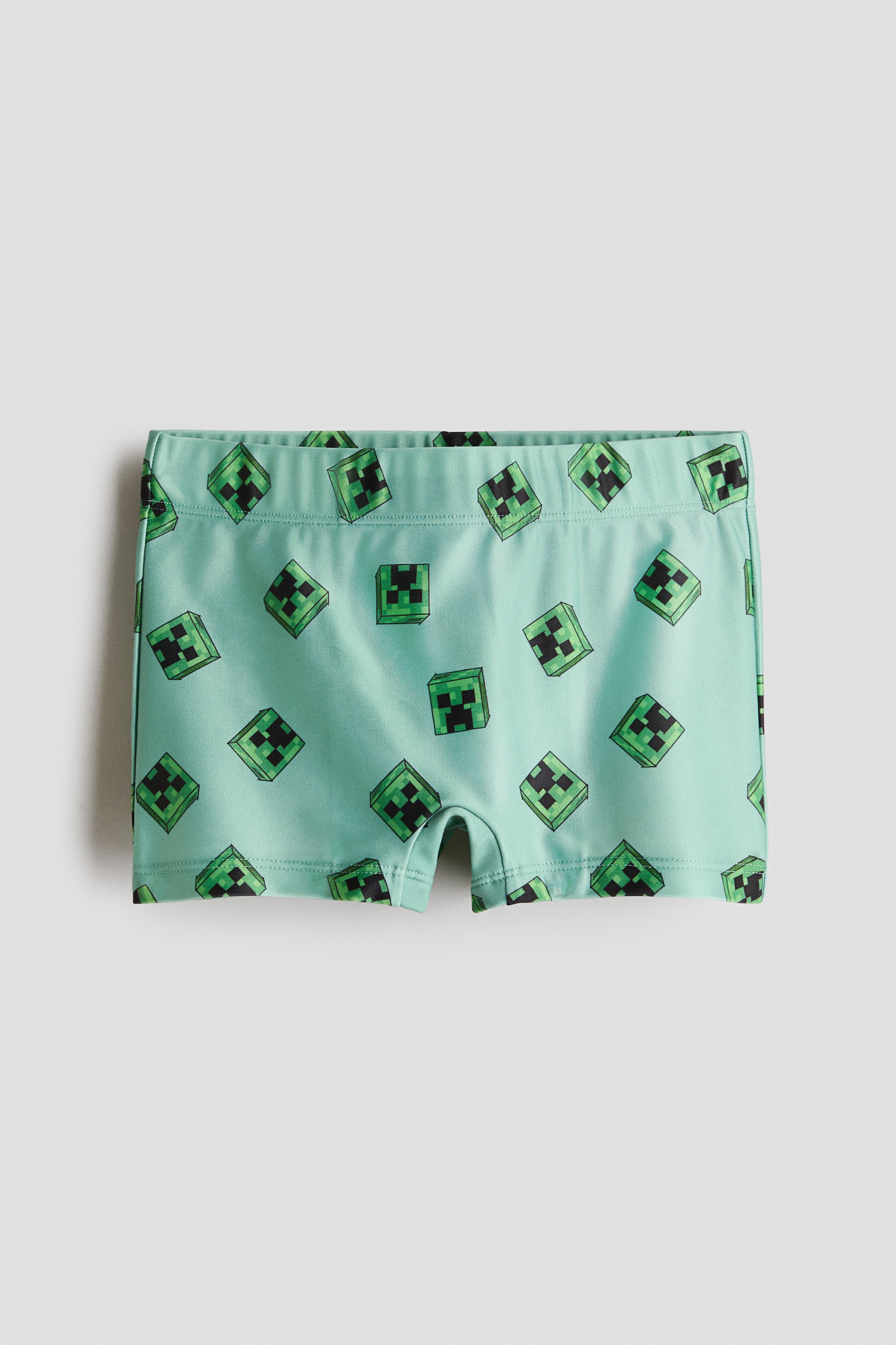 Voir une image plus grande: Trunks de bain imprimés présentés de face, avec une taille élastique et un tissu vert clair recouvert d'un motif répétitif de visages pixelisés verts et noirs.