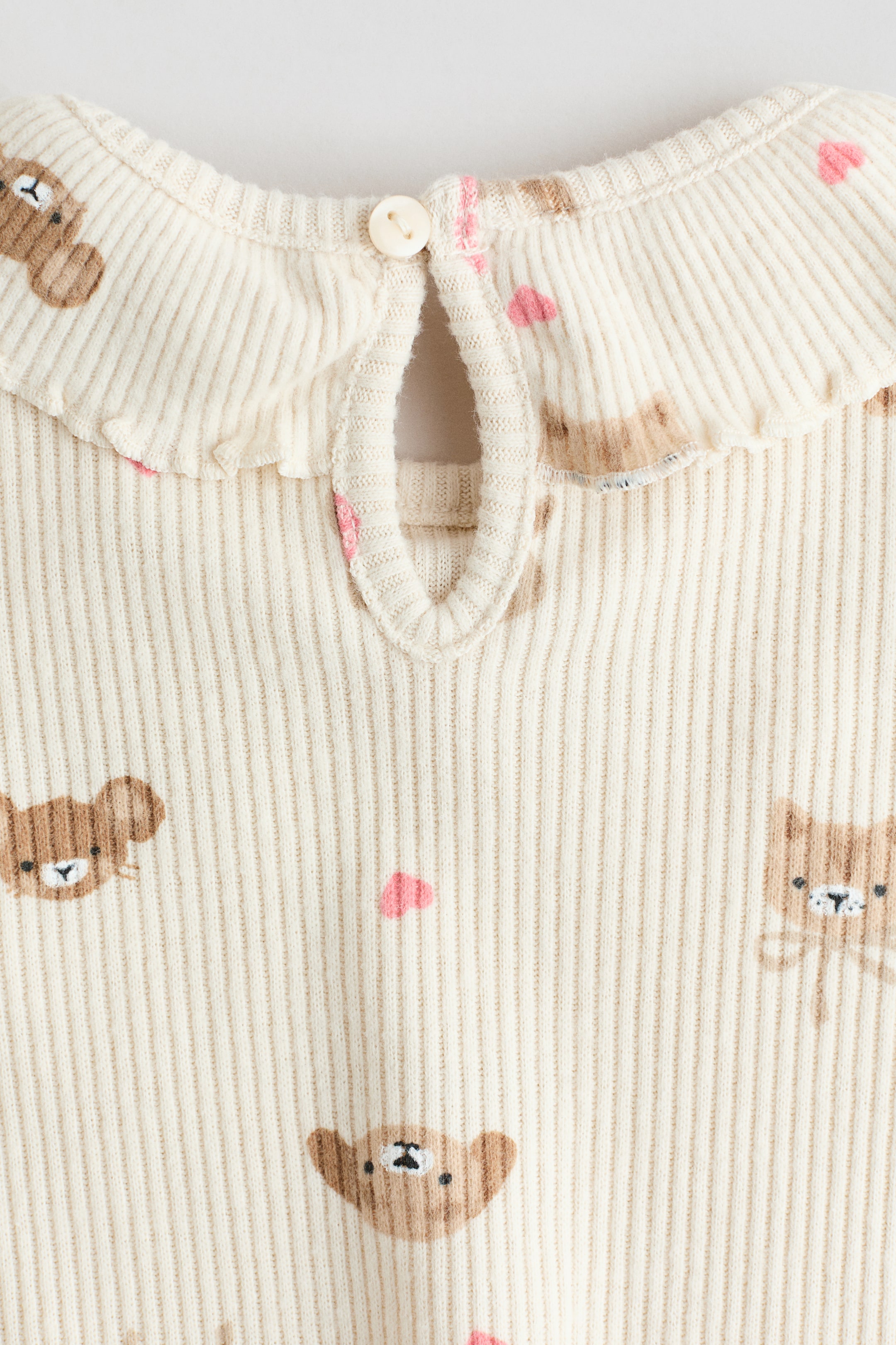 View larger image: A cream ribbed knit garment features a ruffled collar and a keyhole opening at the neck, secured by a cream button. The garment is patterned with repeating brown bear faces and small pink hearts.