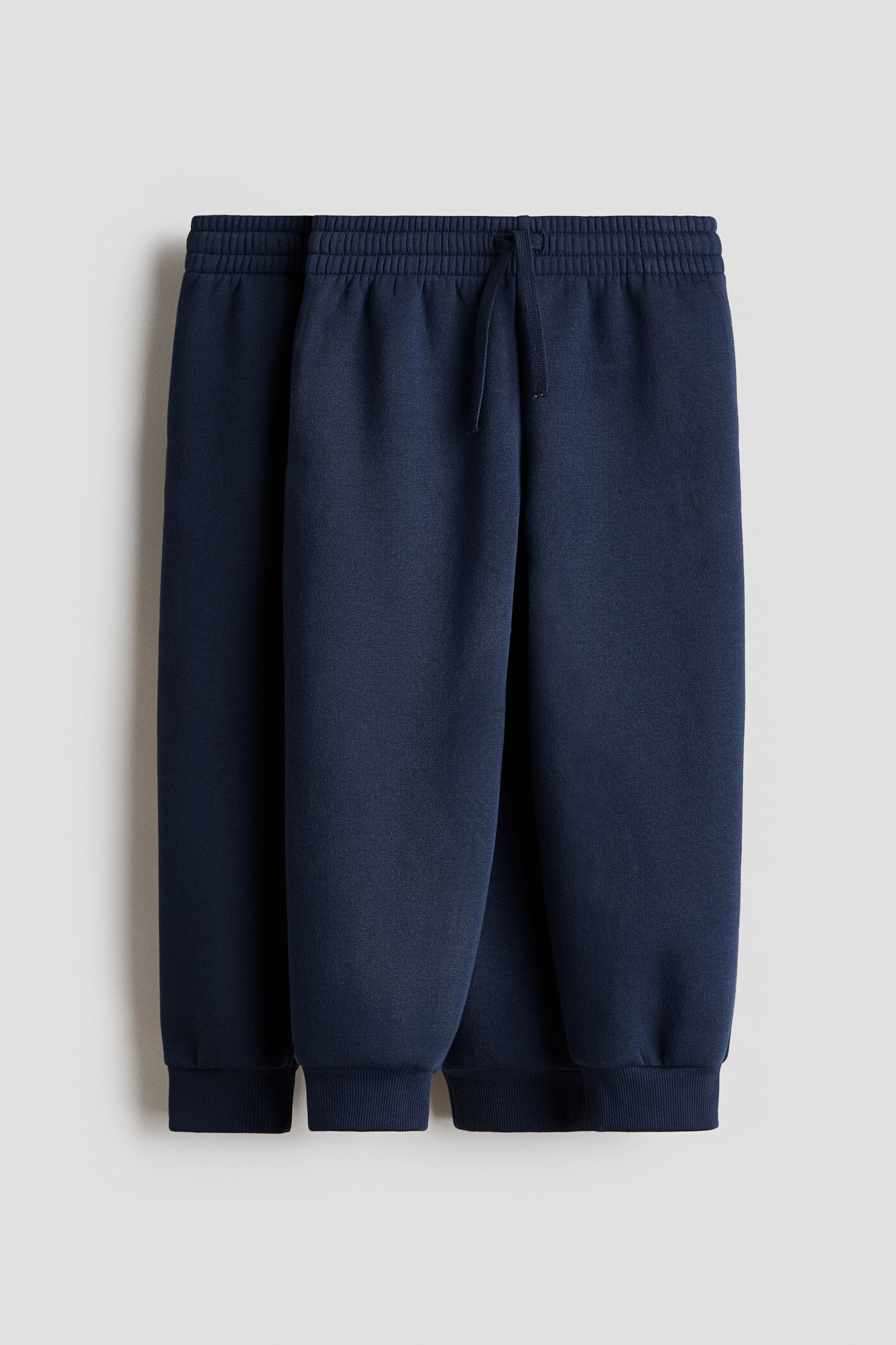 View larger image: Two pairs of navy blue joggers feature elasticated waistbands with a front drawstring tie. The joggers also display ribbed cuffs at the ankles.