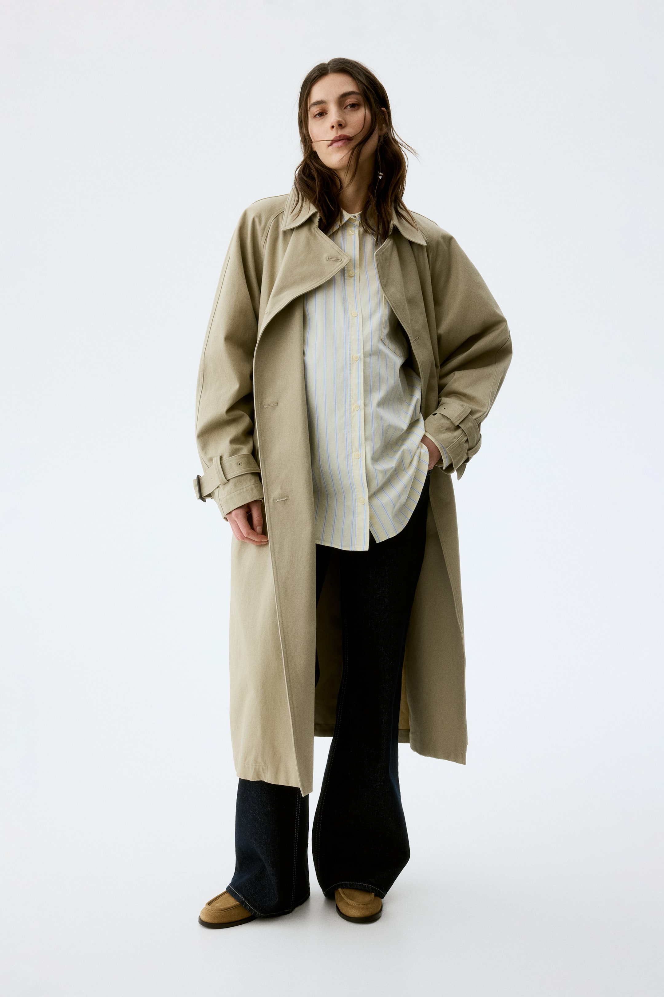 Größeres Bild ansehen: Ein langer, heller khakifarbener Trenchcoat wird präsentiert, nach vorne und leicht nach rechts gerichtet. Er verfügt über einen breiten Kragen, gekerbte Revers, Schultersturmklappen und verstellbare Schnallenmanschetten und wird offen über einem gestreiften Hemd getragen.