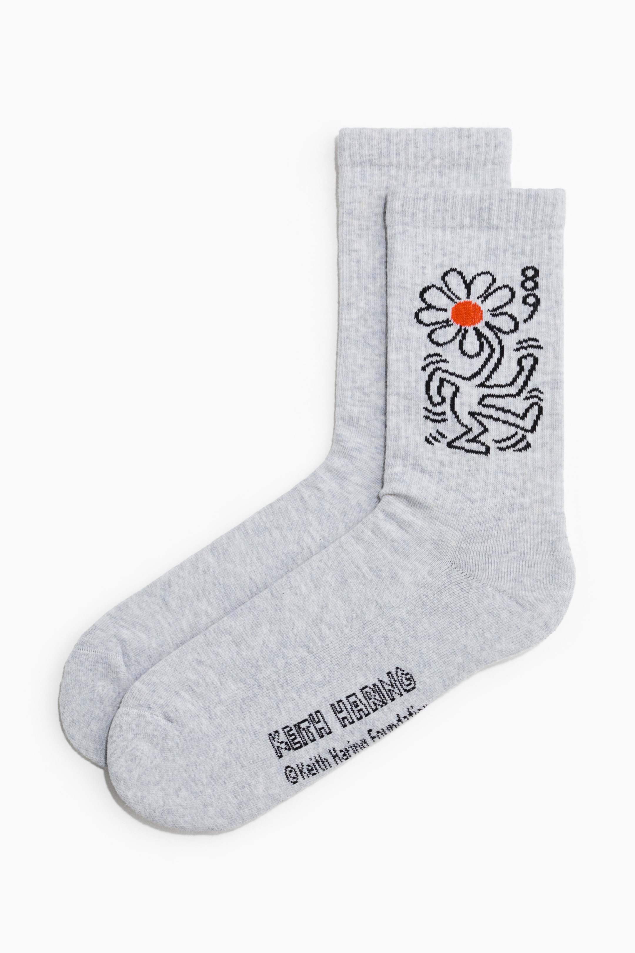 Xem ảnh lớn hơn: Một đôi tất màu xám nhạt, được làm từ chất liệu cotton pha mịn, mềm mại, đang nằm phẳng, đặc trưng với cổ cao, dạng gân. Mặt trước của một chiếc tất hiển thị một họa tiết đường nét màu đen của một hình người cầm một bông hoa có trung tâm màu cam, trong khi khu vực bàn chân có dòng chữ "KEITH HARING" và ""Keith Haring Foundation.""