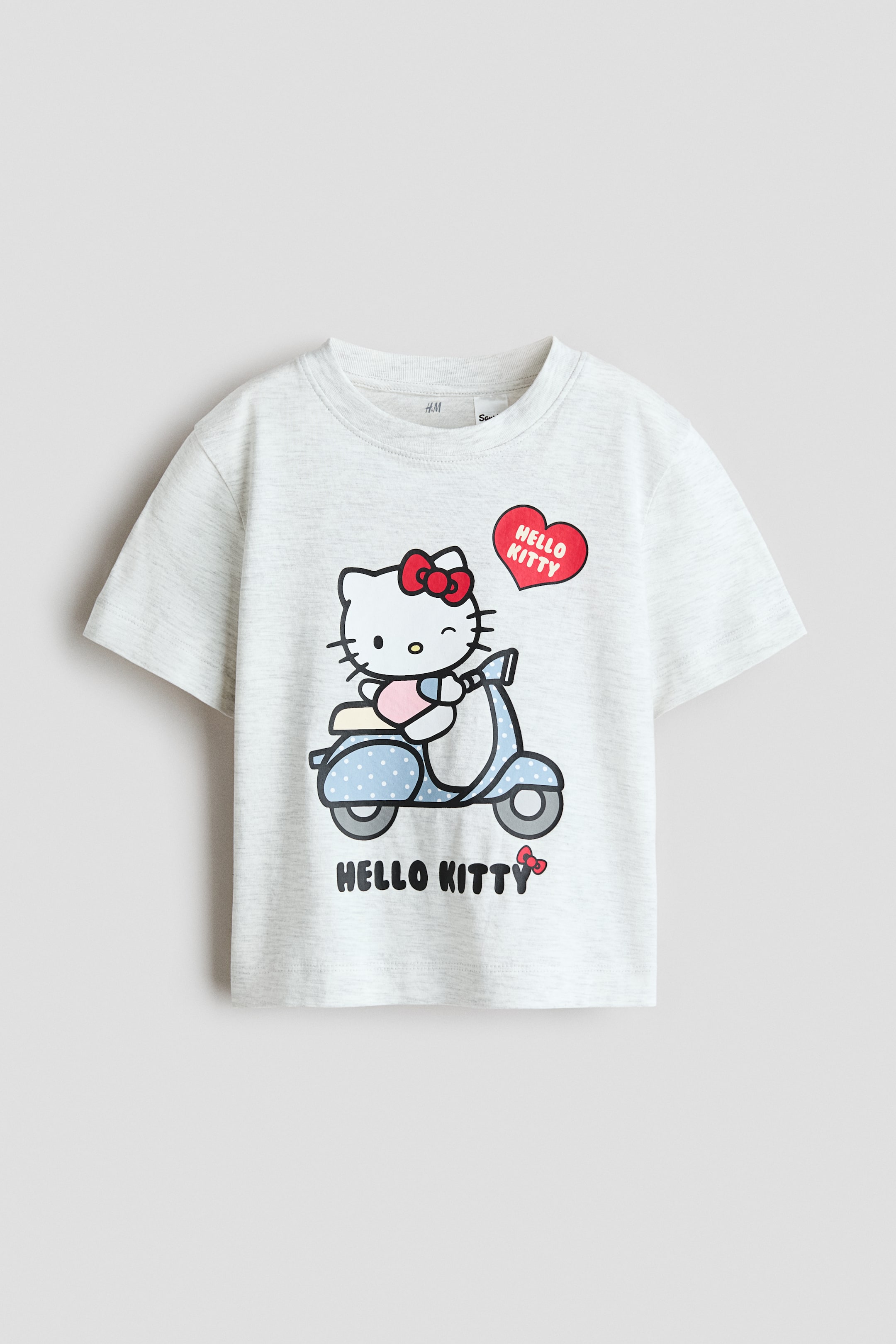 View larger image: A t-shirt is displaying a front view. It is a light grey marl short-sleeved t-shirt with a round neck, featuring a graphic print of Hello Kitty riding a light blue scooter with white polka dots. A red speech bubble above Hello Kitty's head is saying 'HELLO KITTY', and the words 'HELLO KITTY' are also printed below the scooter.