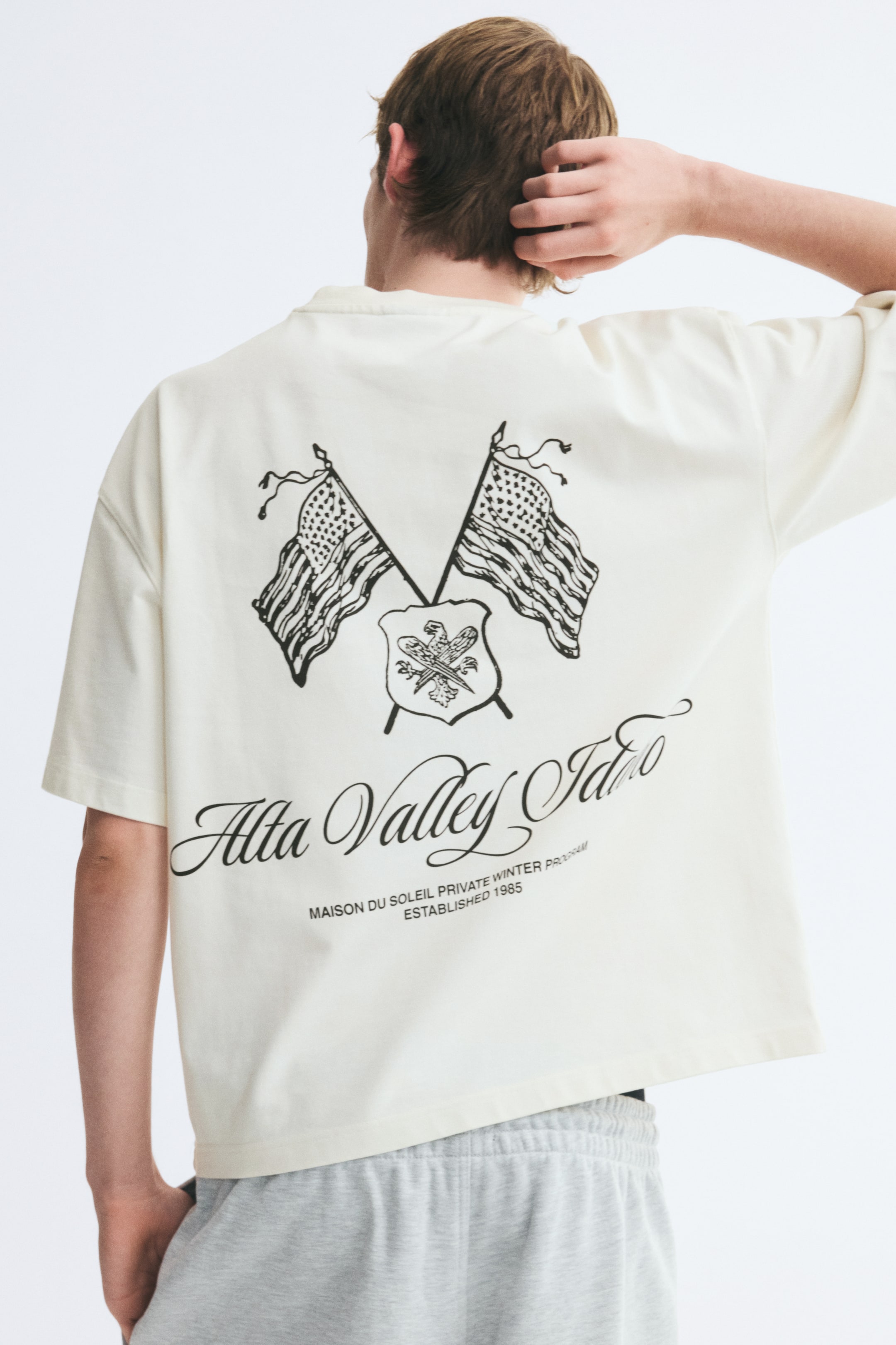 View larger image: An oversized, off-white cotton T-shirt is worn by the model and shown from the back. The T-shirt features dropped shoulders, a straight hem, and a large black graphic print on the back depicting crossed flags, a crest, and cursive text.