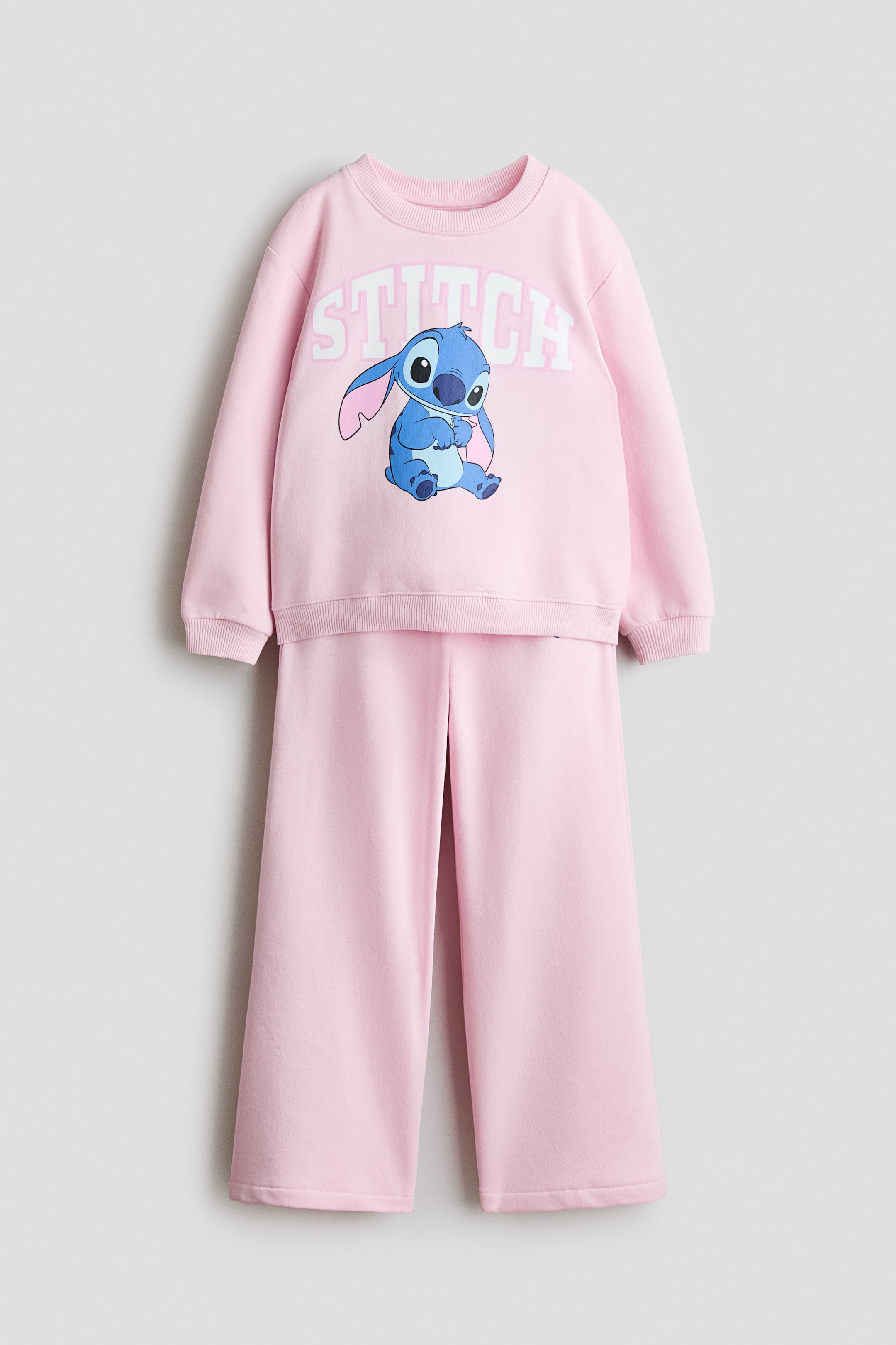 Voir une image plus grande: Un ensemble deux pièces rose, composé d'un sweat-shirt à manches longues et d'un jogger assorti, présente le devant. Le sweat-shirt affiche un imprimé graphique d'un personnage bleu et le texte "STITCH", une encolure ronde bordée de côtes, des épaules tombantes et des bords‑côtes aux poignets et à l'ourlet, tandis que le jogger présente une taille élastiquée et des jambes droites.