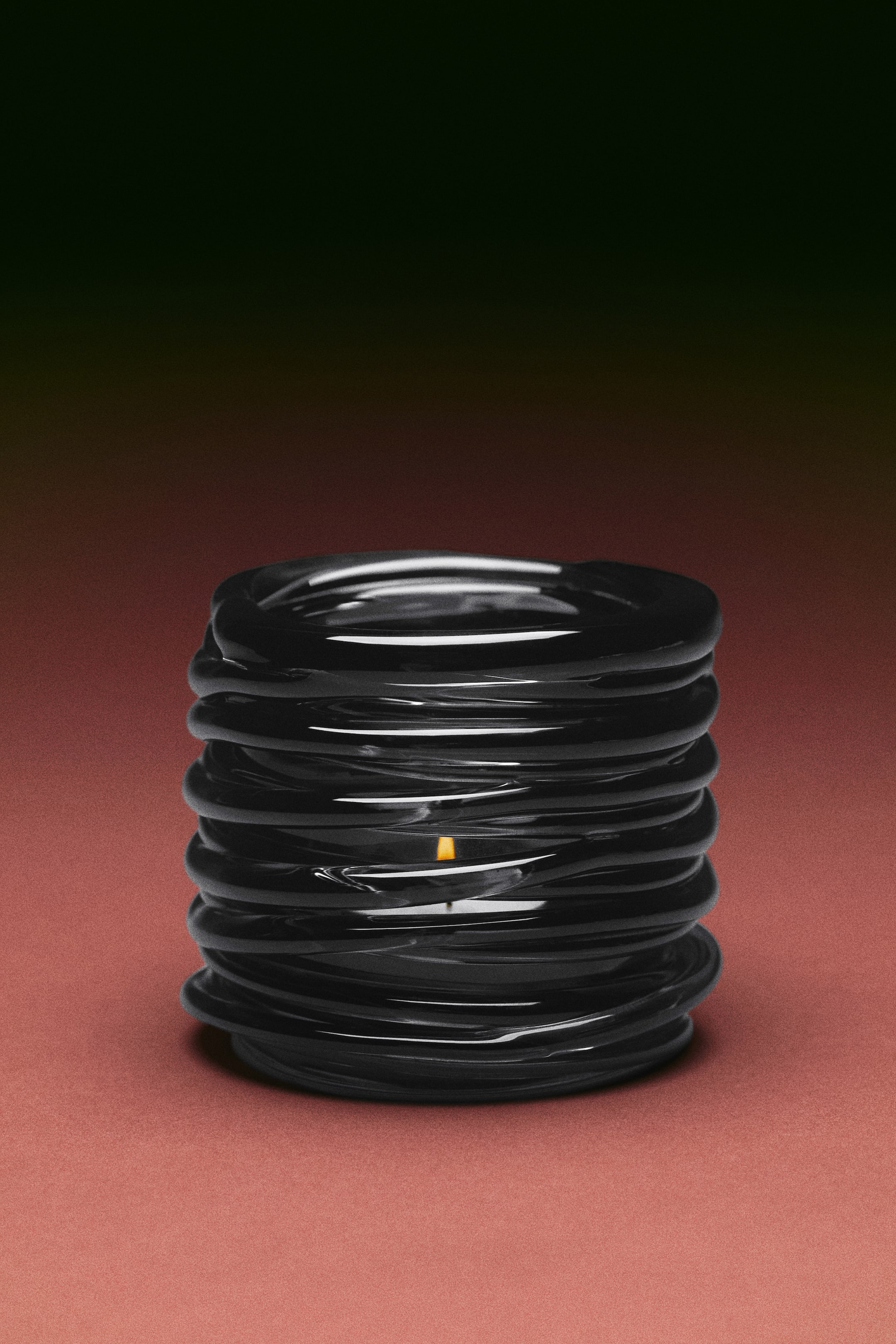 Voir une image plus grande: Un petit bougeoir à chauffe-plat en verre soufflé à la bouche foncé est présenté de face, avec un design sculptural en spirale composé d’anneaux brillants empilés. Une bougie allumée diffuse une douce lueur depuis le centre de la structure.