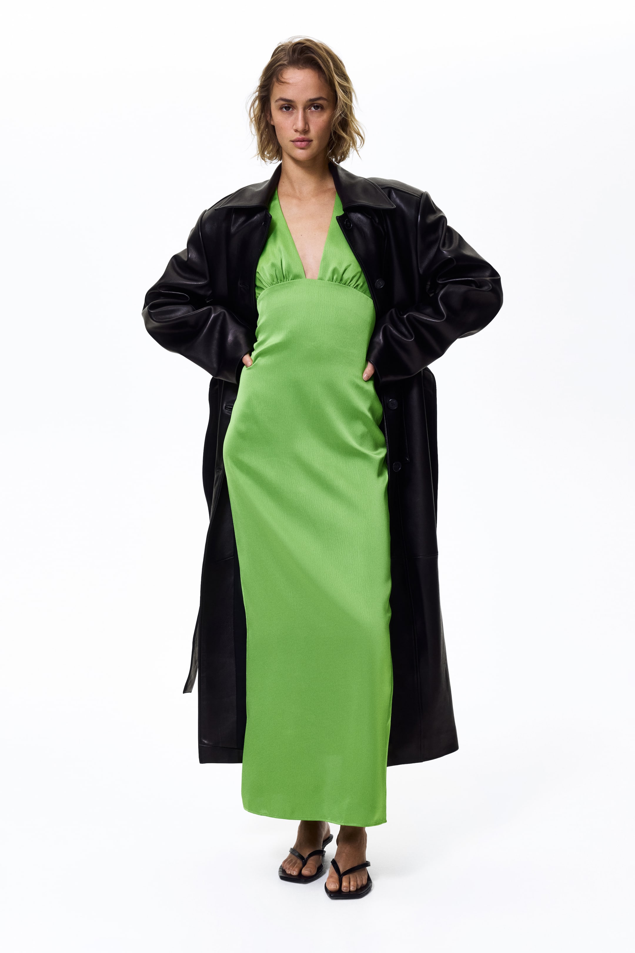 Agrandir l'image: Une robe maxi vert citron à la coupe frontale est présentée, confectionnée dans un tissu subtilement texturé. Elle arbore un décolleté en V et un détail froncé au niveau du buste, portée sous un trench-coat noir ouvert et stylisée avec des sandales noires.