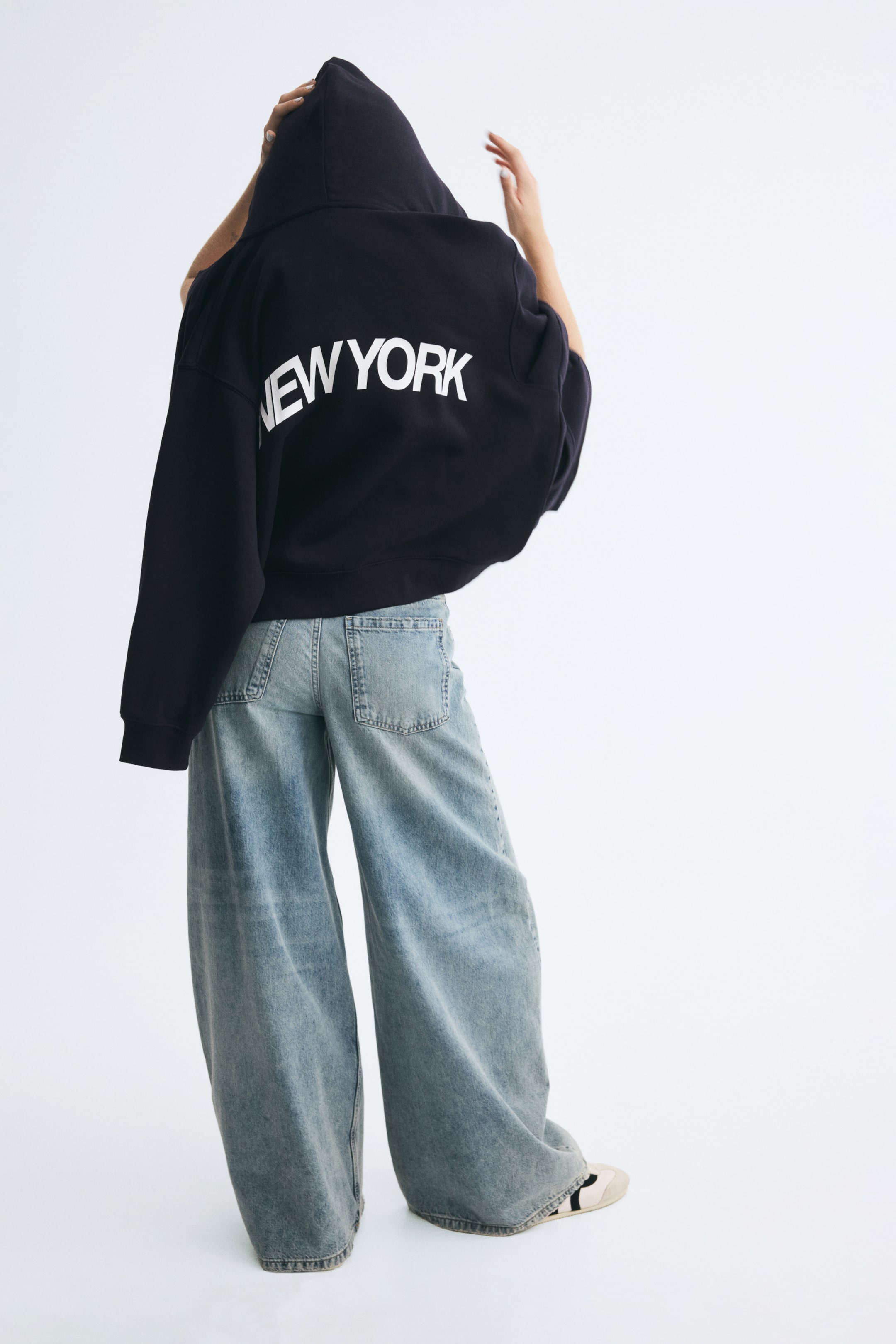 View larger image: A black zip-up hoodie with "NEW YORK" printed in white arched text across the back is being worn by the model, who is facing away from the camera with the hood up. The model is adjusting the hood with both hands, styling the garment with light-wash, wide-leg denim jeans.