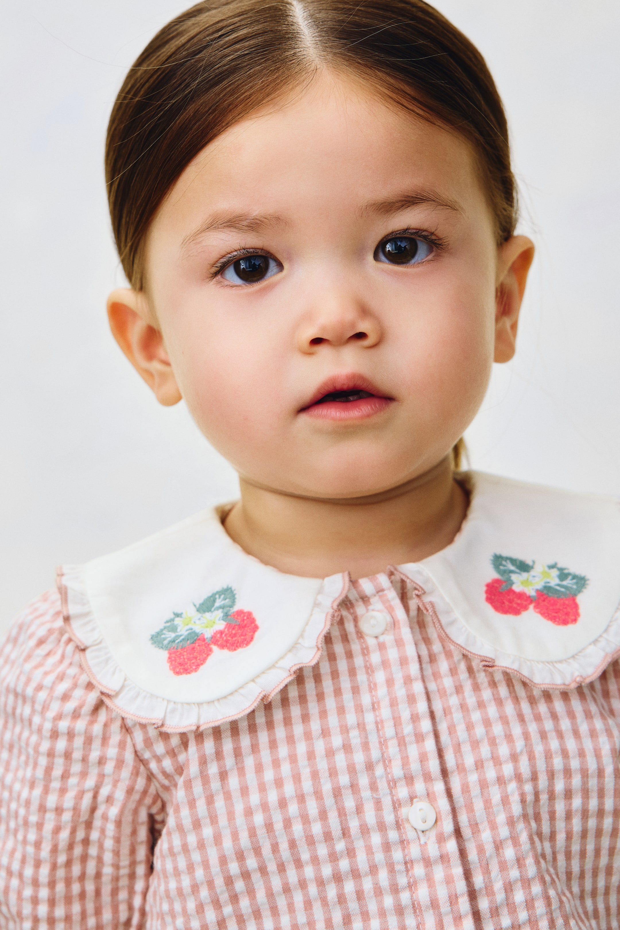 Größeres Bild ansehen: Ein rosa-weißes Vichy-Karo-Oberteil mit einem abgerundeten weißen Kragen, der mit gestickten Erdbeeren und einem gerüschten Rand verziert ist. Der Verschluss vorne wird mit weißen Knöpfen geschlossen.