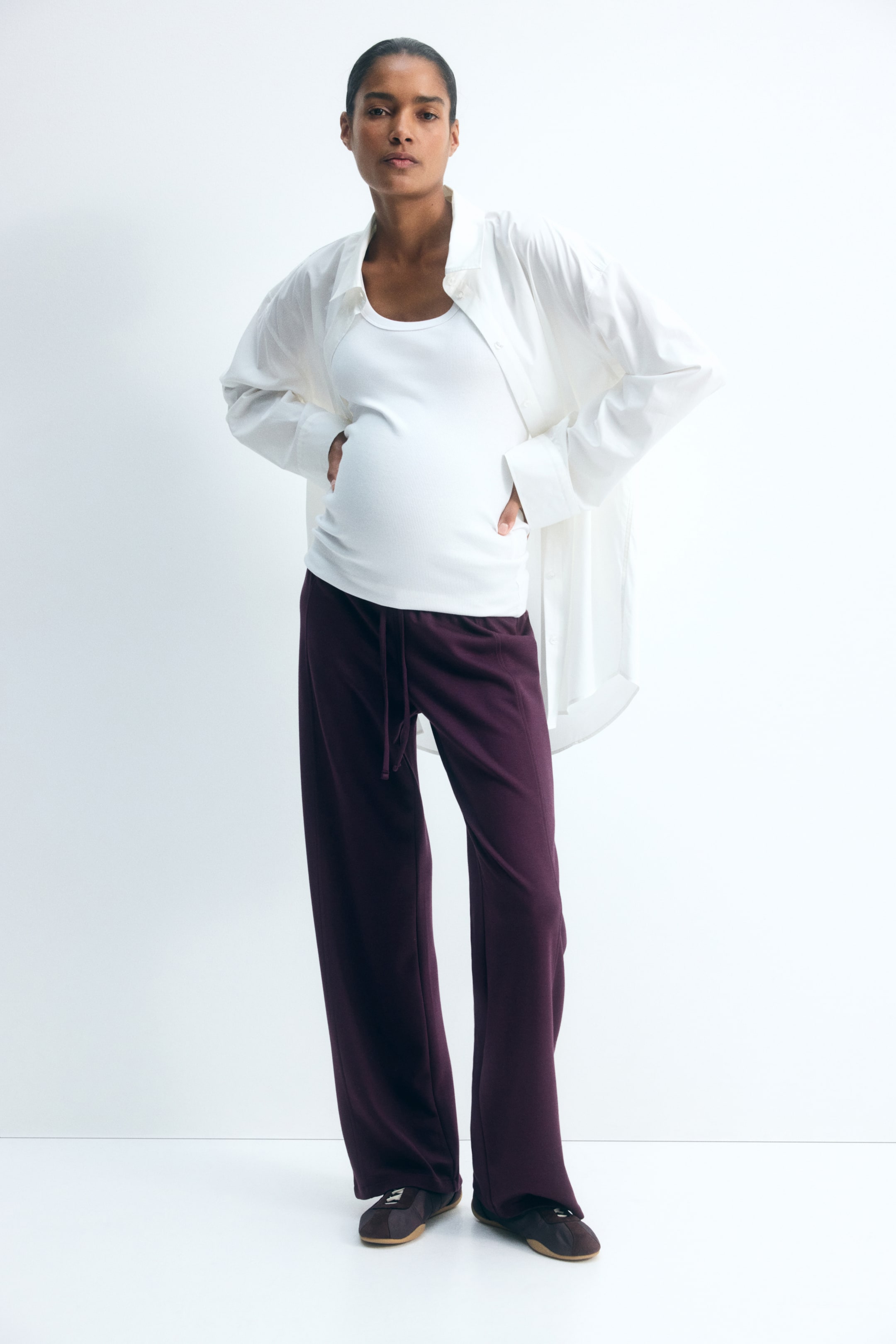 View larger image: Dark purple wide-leg trousers featuring a visible front seam and a narrow drawstring waist are worn by the model. The relaxed-fit trousers are styled with a white tank top and an open white button-up shirt, highlighting the low, elasticated waistline.