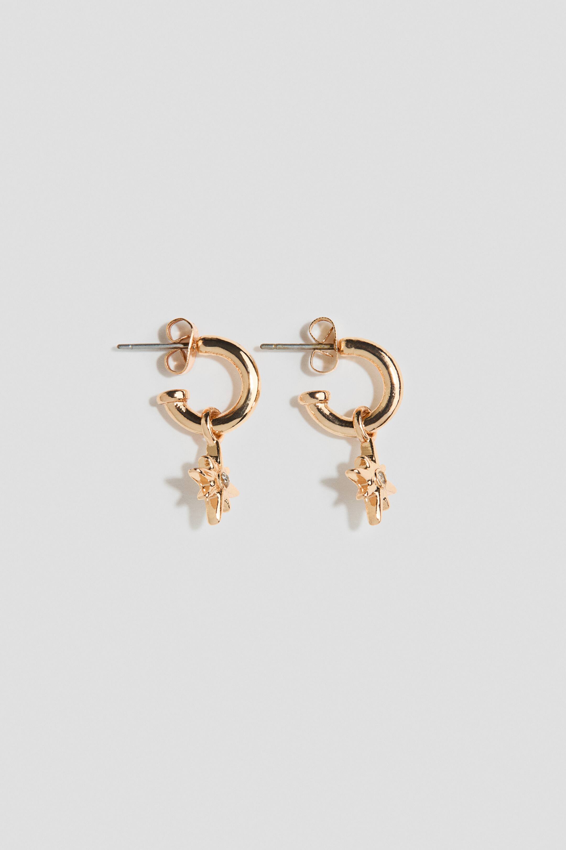 View larger image: A pair of gold-toned hoop earrings is shown front-facing, featuring small, polished hoops with post-back closures. Each hoop supports a dangling starburst-shaped charm accented with a small, clear gemstone at the center.