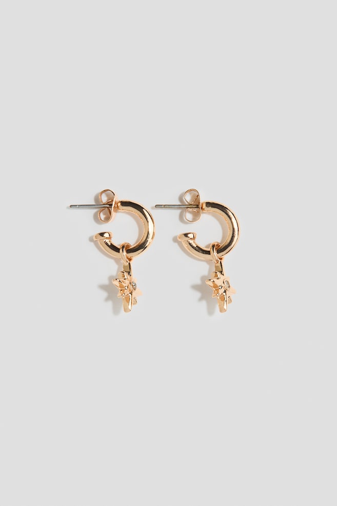 A pair of gold-toned hoop earrings is shown facing front, featuring small, polished hoops with post-back closures. Each hoop is supporting a dangling starburst-shaped charm embellished with a small, clear gemstone in the center. - 1