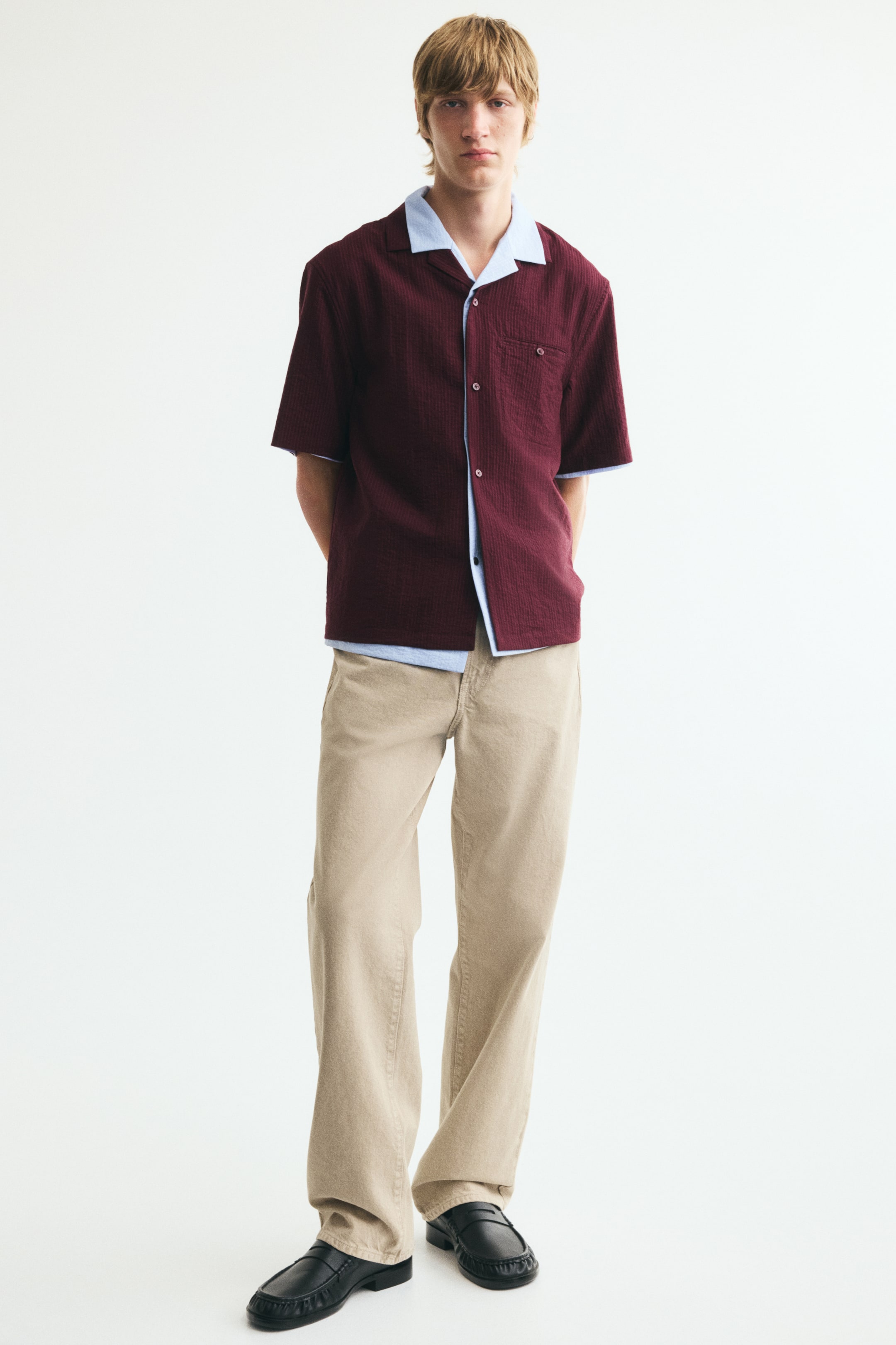 View larger image: A burgundy short-sleeved seersucker shirt with light blue trim, featuring a resort collar, French front, an open chest pocket with a button-and-loop fastener, and a straight-cut hem, is being worn by the model unbuttoned over a light blue undershirt, showcasing its relaxed fit.