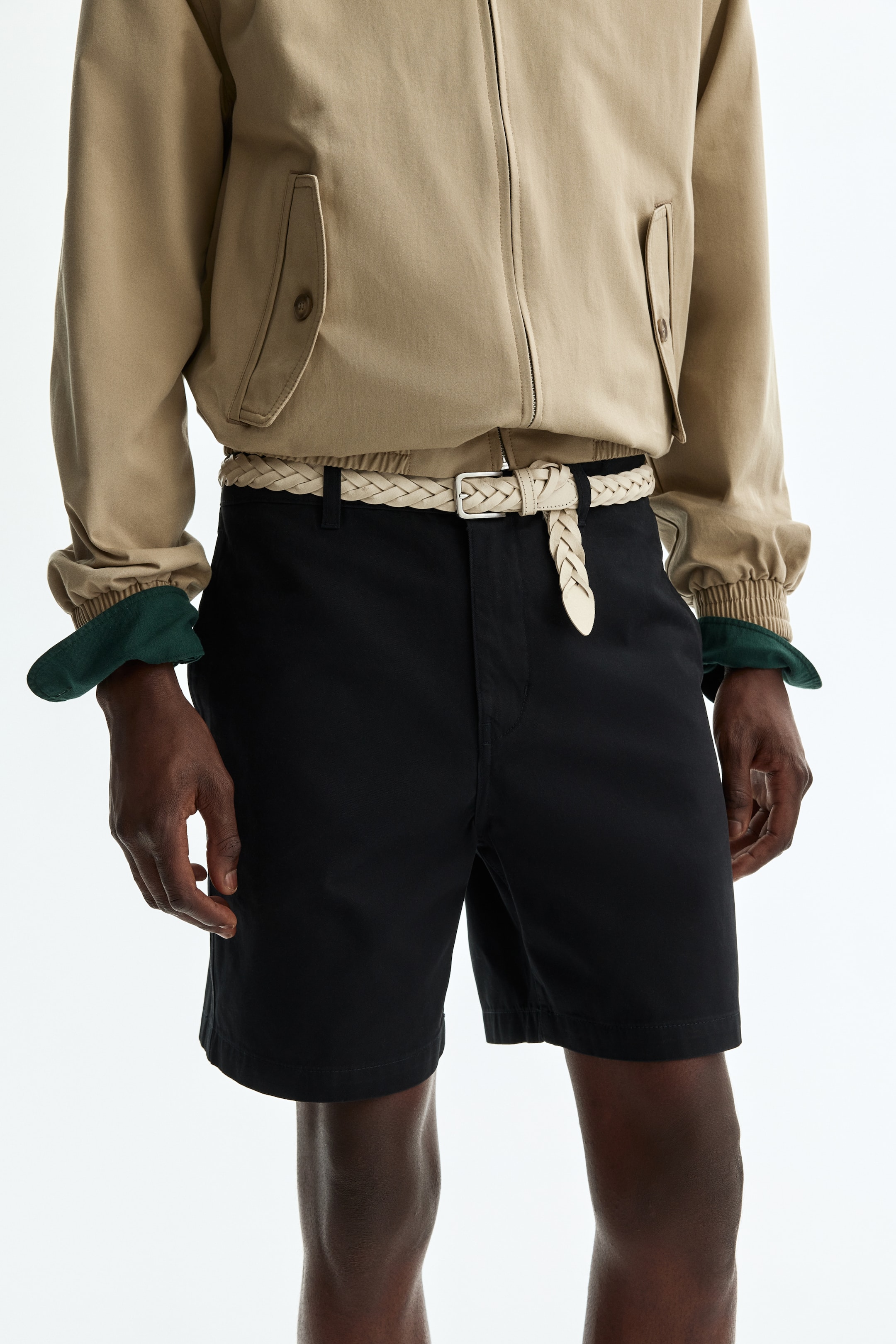View larger image: The model is wearing black chino shorts crafted from cotton twill. They have a zip fly with a button closure, side pockets, and a relaxed fit, complemented by a braided belt threaded through the belt loops.