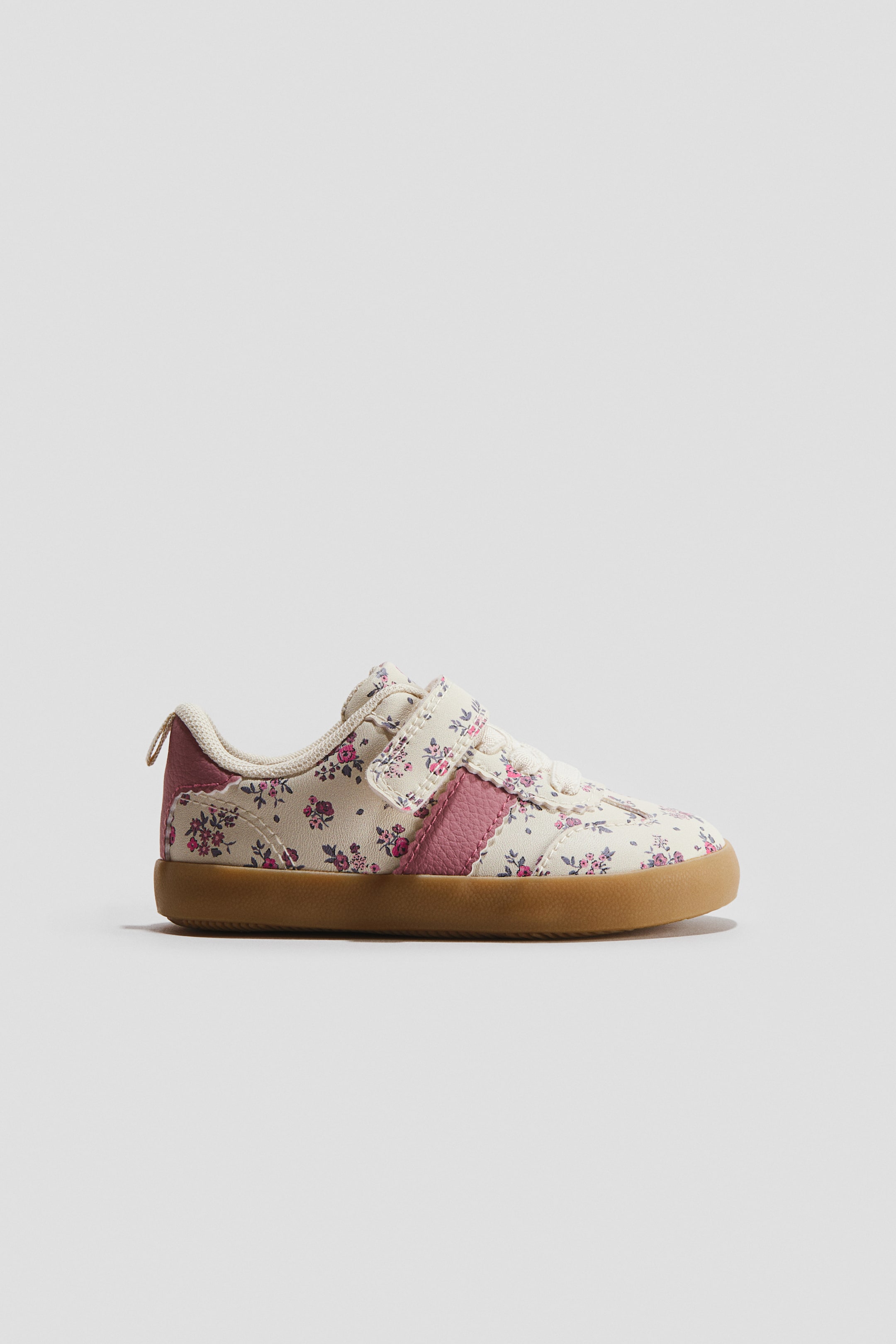 Ver imagen más grande: Las zapatillas minimalistas presentan una parte superior en color crema con un estampado floral en tonos rosa y verde, vistas desde un ángulo lateral. Cuentan con un cierre de correa de velcro, una franja lateral acanalada en rosa y una suela plana de goma marrón.