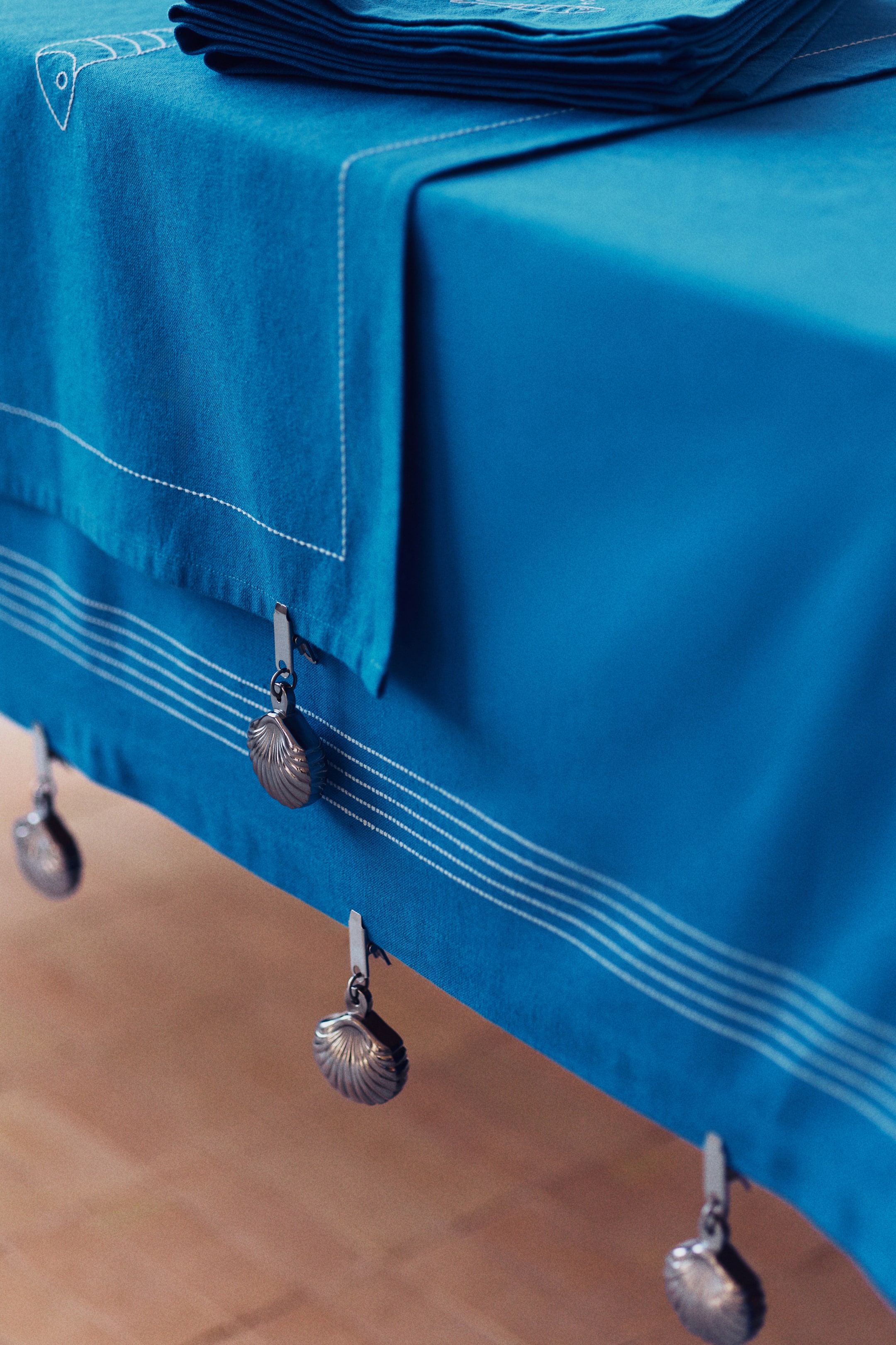 View larger image: A blue tablecloth covers a table, with a blue table runner draped across it, featuring white dashed stitching and a white fish embroidery. Silver-toned, shell-shaped table weights hang from clips along the tablecloth's bottom edge, which also displays thin white horizontal lines.