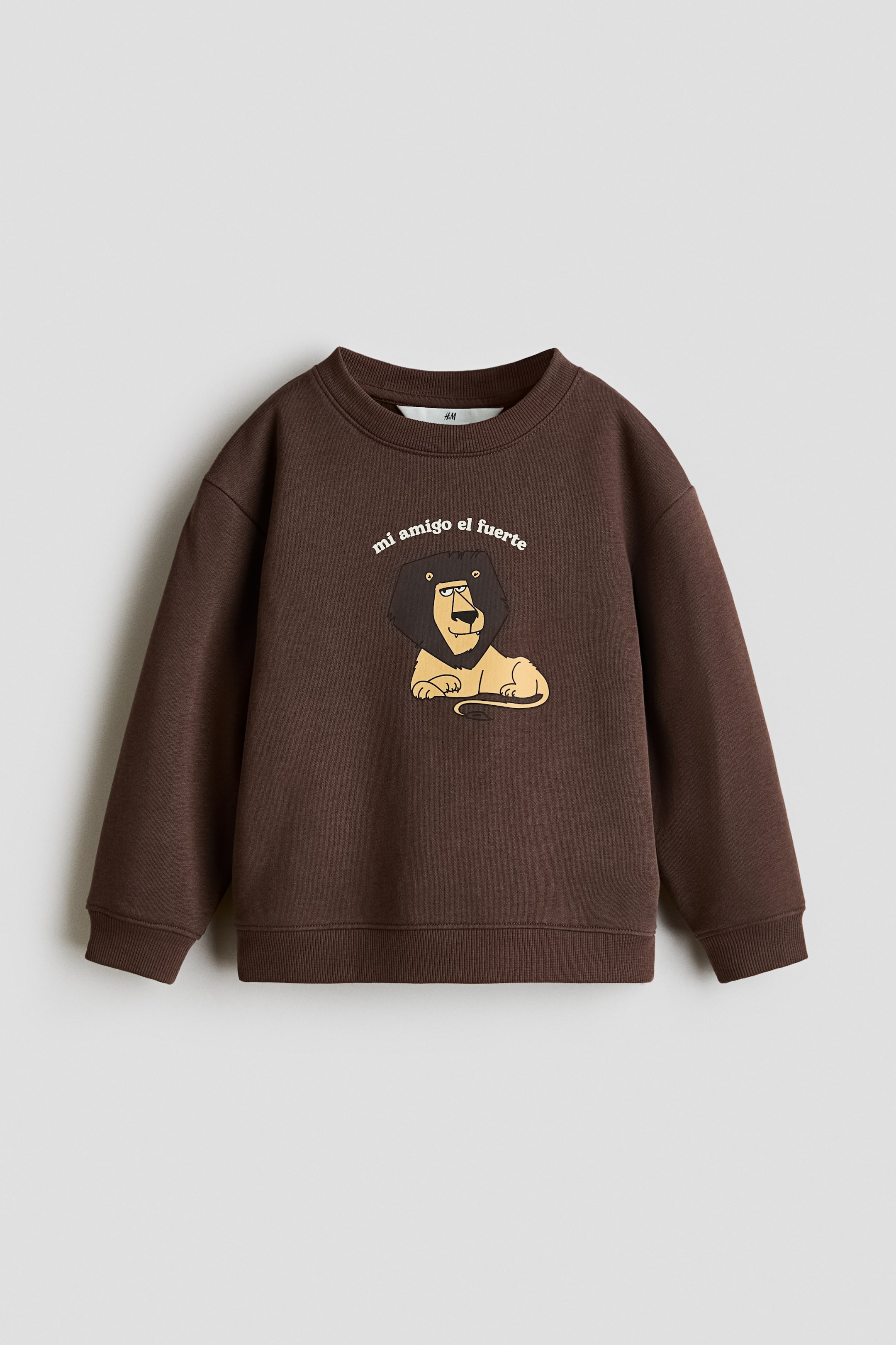 View larger image: A brown sweatshirt is laid flat, featuring a ribbed crew neck, ribbed cuffs, and a ribbed hem. The front is printed with a cartoon lion graphic and the curved text "mi amigo el fuerte" above it.