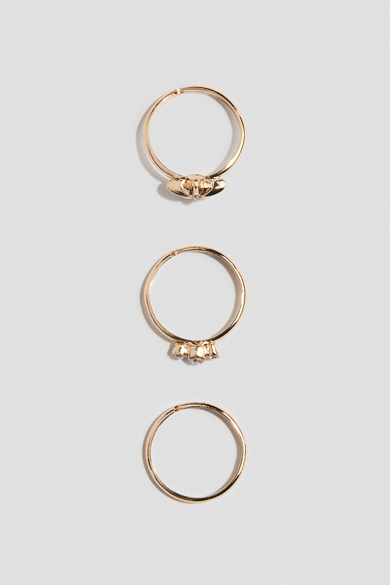 Tres anillos en tono dorado se muestran en vertical para destacar sus diseños desde un ángulo frontal. El anillo superior presenta un pequeño adorno en forma de lazo con una diminuta piedra transparente, el anillo central muestra un grupo de tres piedras de strass transparentes, y el anillo inferior presenta una banda lisa y sin adornos.