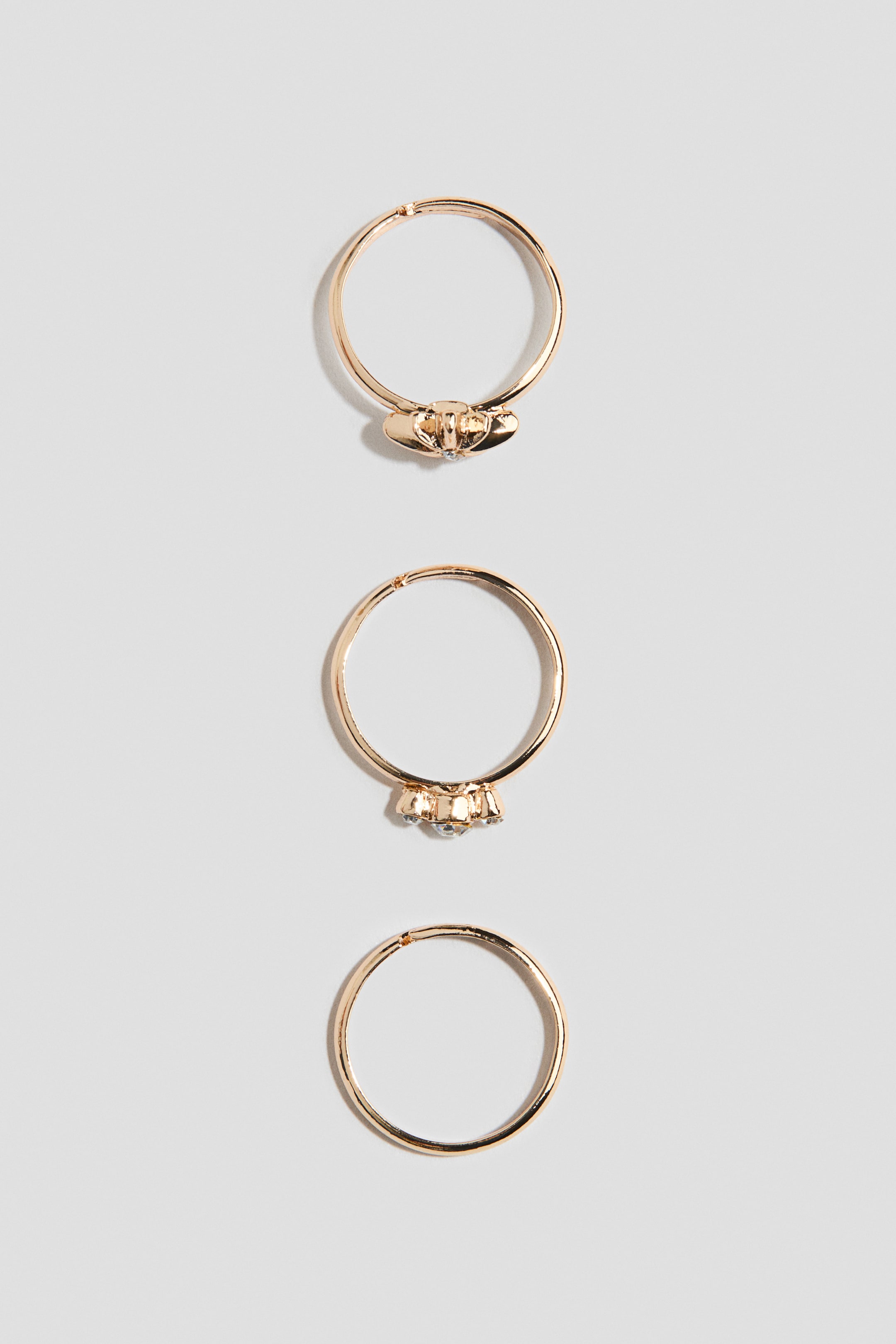 Ver imagen más grande: Tres anillos en tono dorado se muestran en vertical para destacar sus diseños desde un ángulo frontal. El anillo superior presenta un pequeño adorno en forma de lazo con una diminuta piedra transparente, el anillo central muestra un grupo de tres piedras de strass transparentes, y el anillo inferior presenta una banda lisa y sin adornos.