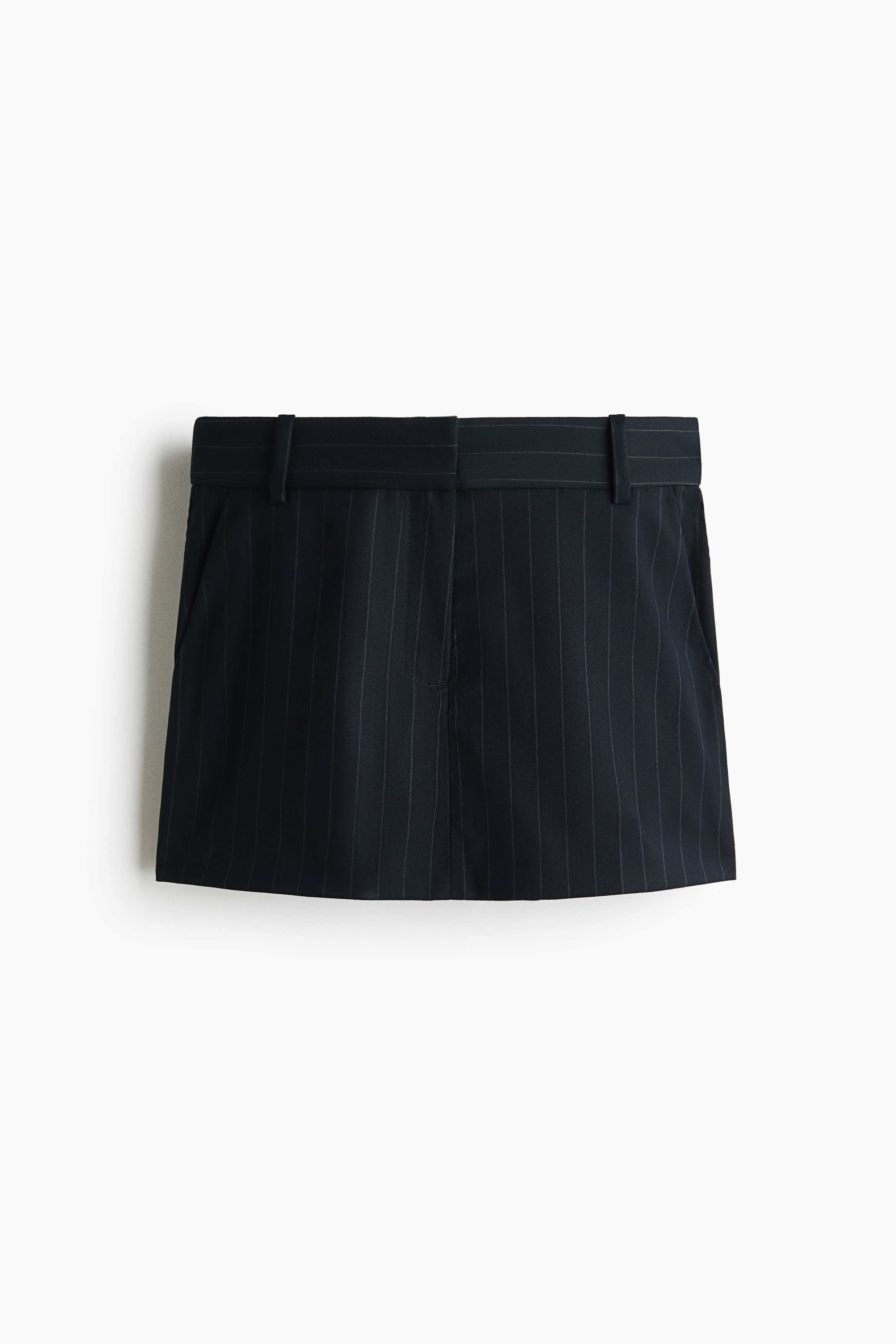 La modelo lleva puesto un skort negro con rayas diplomÃ¡ticas, de cintura regular con trabillas para cinturÃ³n y un bolsillo lateral diagonal. El skort se complementa con una blusa sin mangas a juego con rayas diplomÃ¡ticas y un suÃ©ter rojo brillante colocado sobre los hombros.