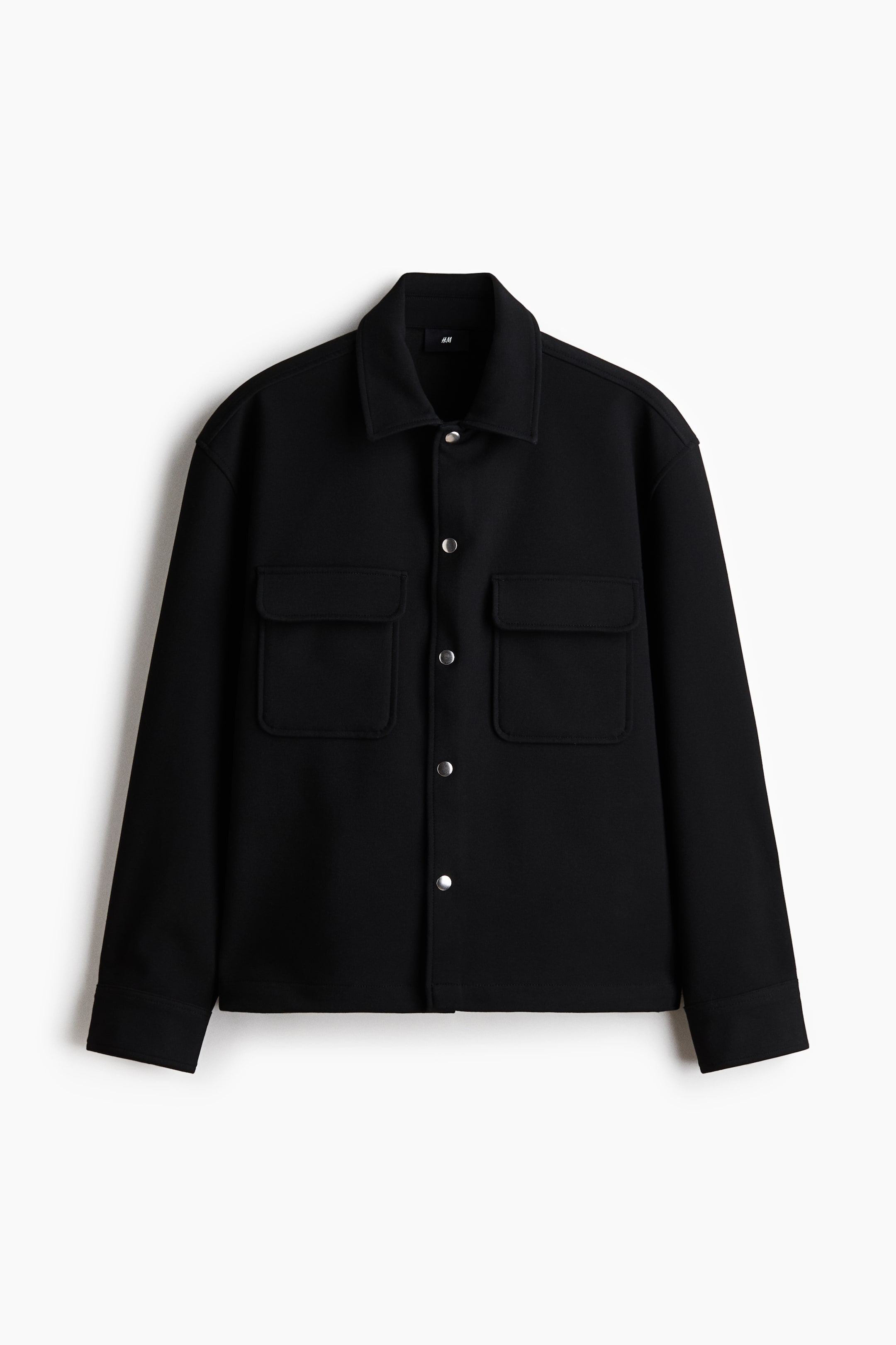 Ver imagen más grande: Una chaqueta negra estructurada está orientada hacia el frente, con un cuello clásico y botones automáticos de tono plateado en la parte delantera. Presenta dos bolsillos de parche con solapa en el pecho y mangas largas con puños.