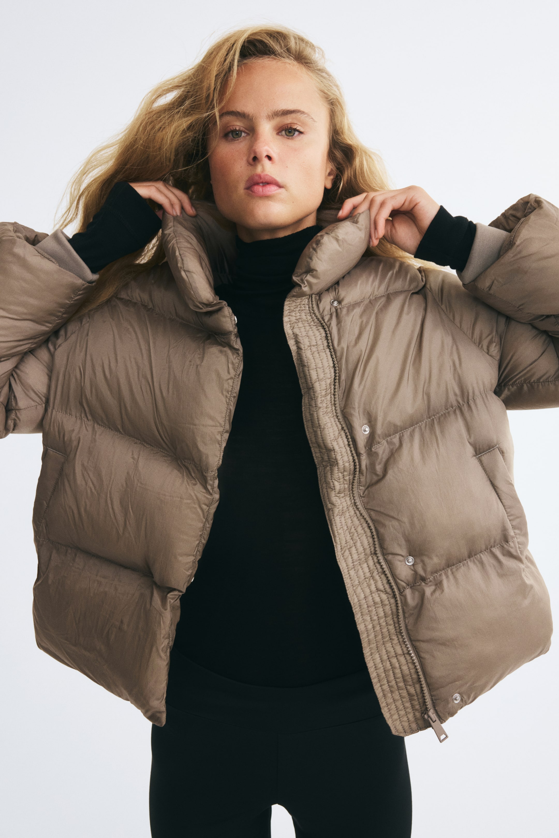 Voir une image plus grande: Une veste matelassée surdimensionnée taupe, en tissage de nylon matelassé, est portée par le modèle, qui tient le col cheminée montant des deux mains. La veste est portée ouverte sur un col roulé noir, mettant en valeur sa fermeture éclair à l’avant, sa patte coupe-vent à boutons-pression et ses poches passepoilées à l’avant.