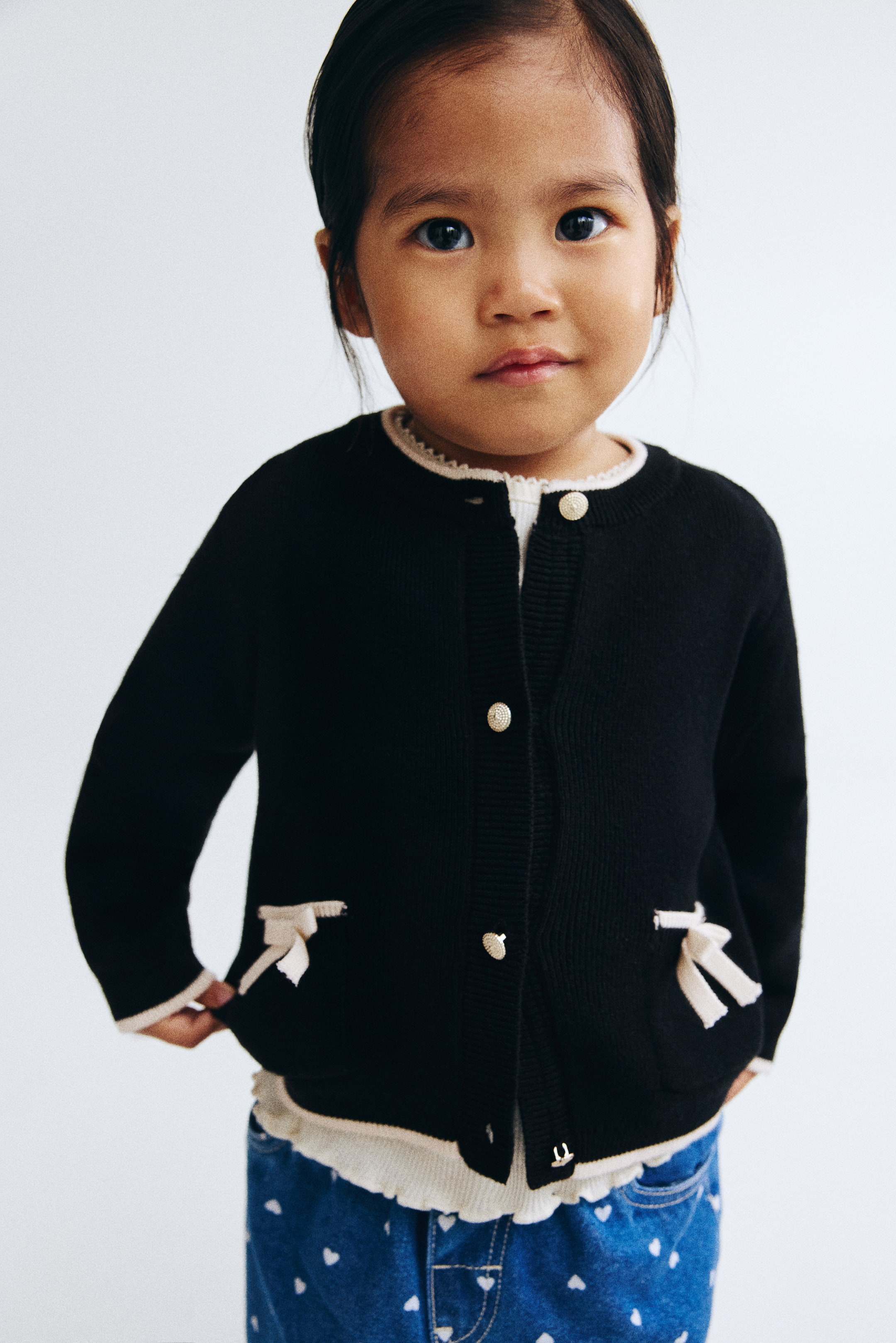 View larger image: A black, soft, fine-knit cotton cardigan is showcased from the front, featuring a round neckline with light-colored ribbed trim and gold-toned heart-shaped metal buttons running down the front. It includes two patch pockets with light-colored trim, each embellished with a light-colored bow, and is completed with light-colored ribbed cuffs and hem.