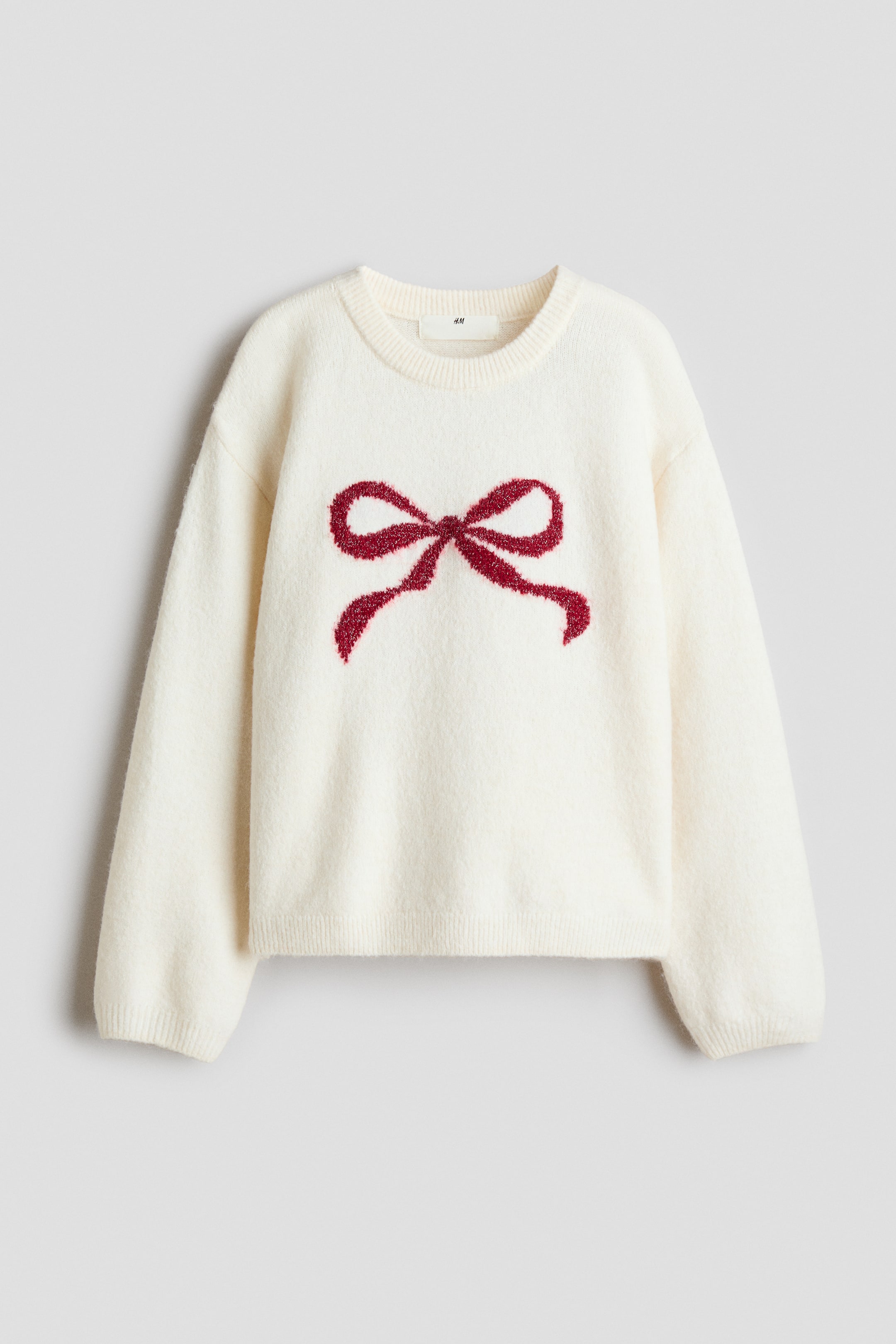 Agrandir l'image: Un pull en maille écru à coupe ample est présenté de face, mettant en valeur un motif de nœud texturé rouge au centre de la poitrine. Le pull présente un col rond côtelé, des épaules tombantes et des finitions côtelées aux poignets et à l’ourlet.