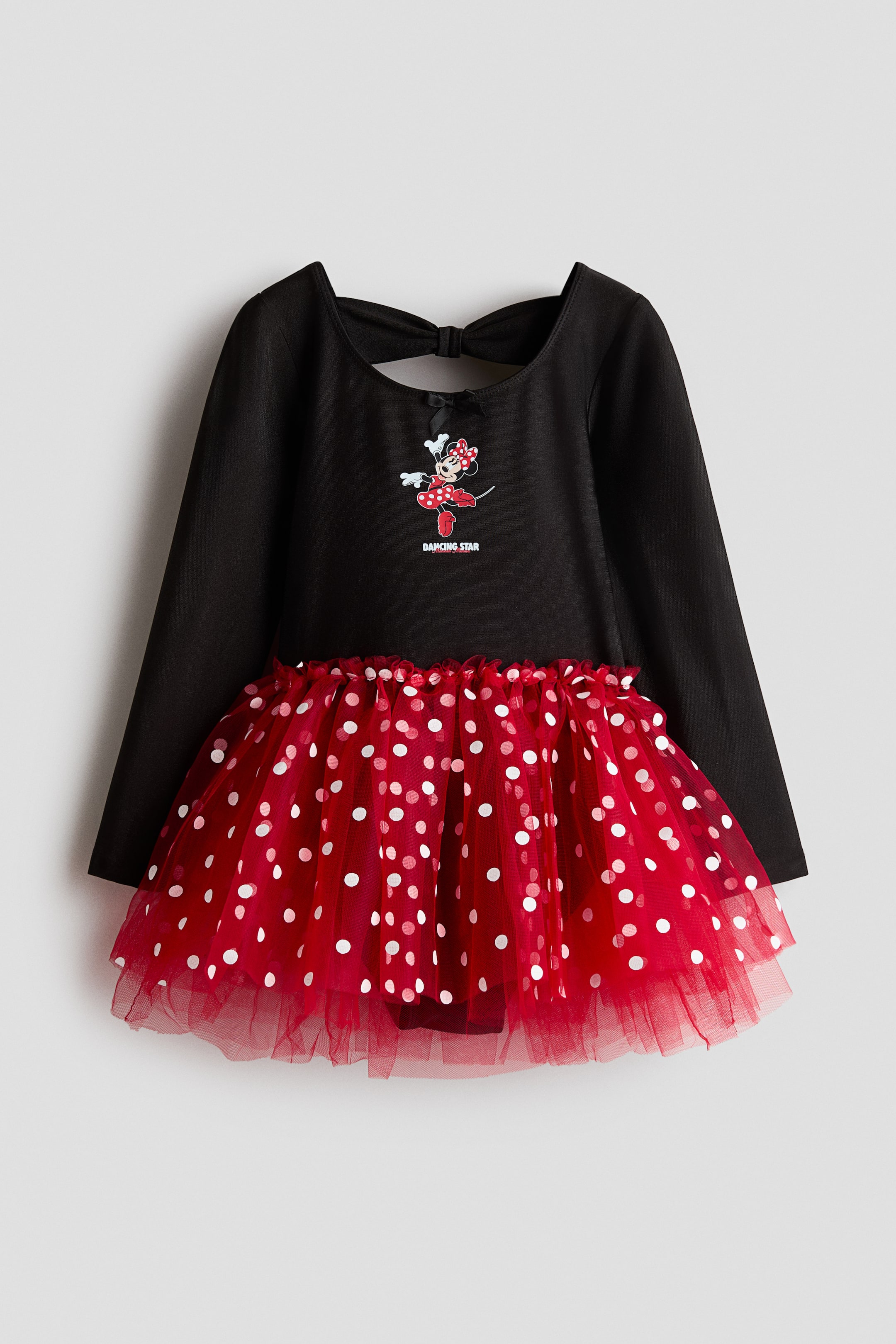 View larger image: A child’s dance dress is shown from the front, featuring a long-sleeved black bodice and a flared, layered red tulle skirt with white polka dots. The bodice features a small black bow at the round neckline, a central graphic print of Minnie Mouse with ‘DANCING STAR’ text, and a bow-shaped cut-out detail at the back.