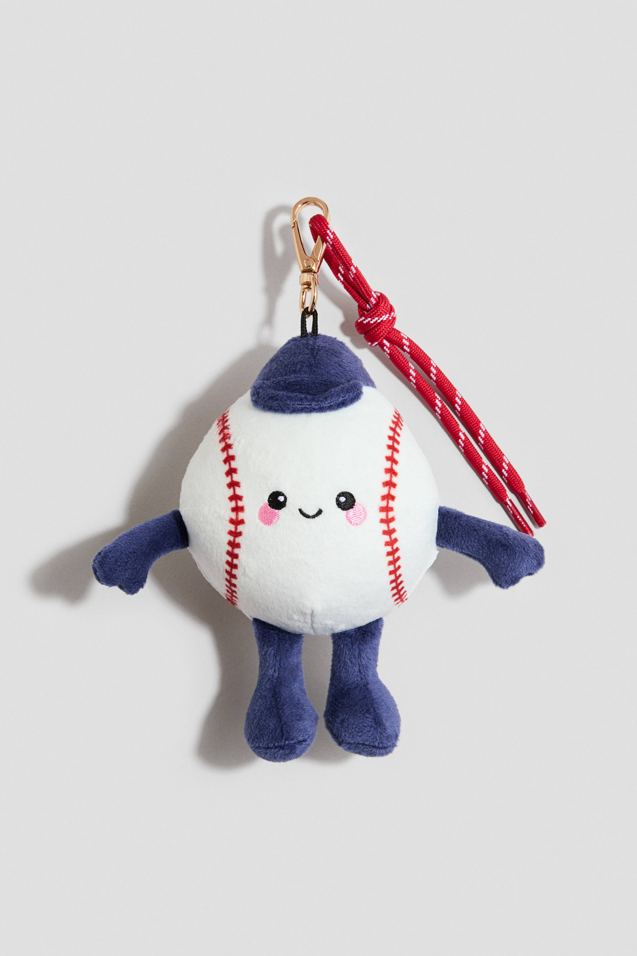 Ampliar la imagen: Un amuleto de bolso presenta un gran juguete de peluche acolchado, mirando hacia ADELANTE, diseñado como una pelota de béisbol blanca con costuras rojas, pequeños brazos y piernas azul oscuro, y una gorra azul oscuro en su cabeza. El juguete muestra una cara bordada con ojos ovalados negros, una pequeña sonrisa y un rubor rosado, mientras que un gancho mosquetón de metal dorado se adhiere a la parte superior de la gorra, y una correa trenzada roja y blanca en espiral también se conecta al gancho.