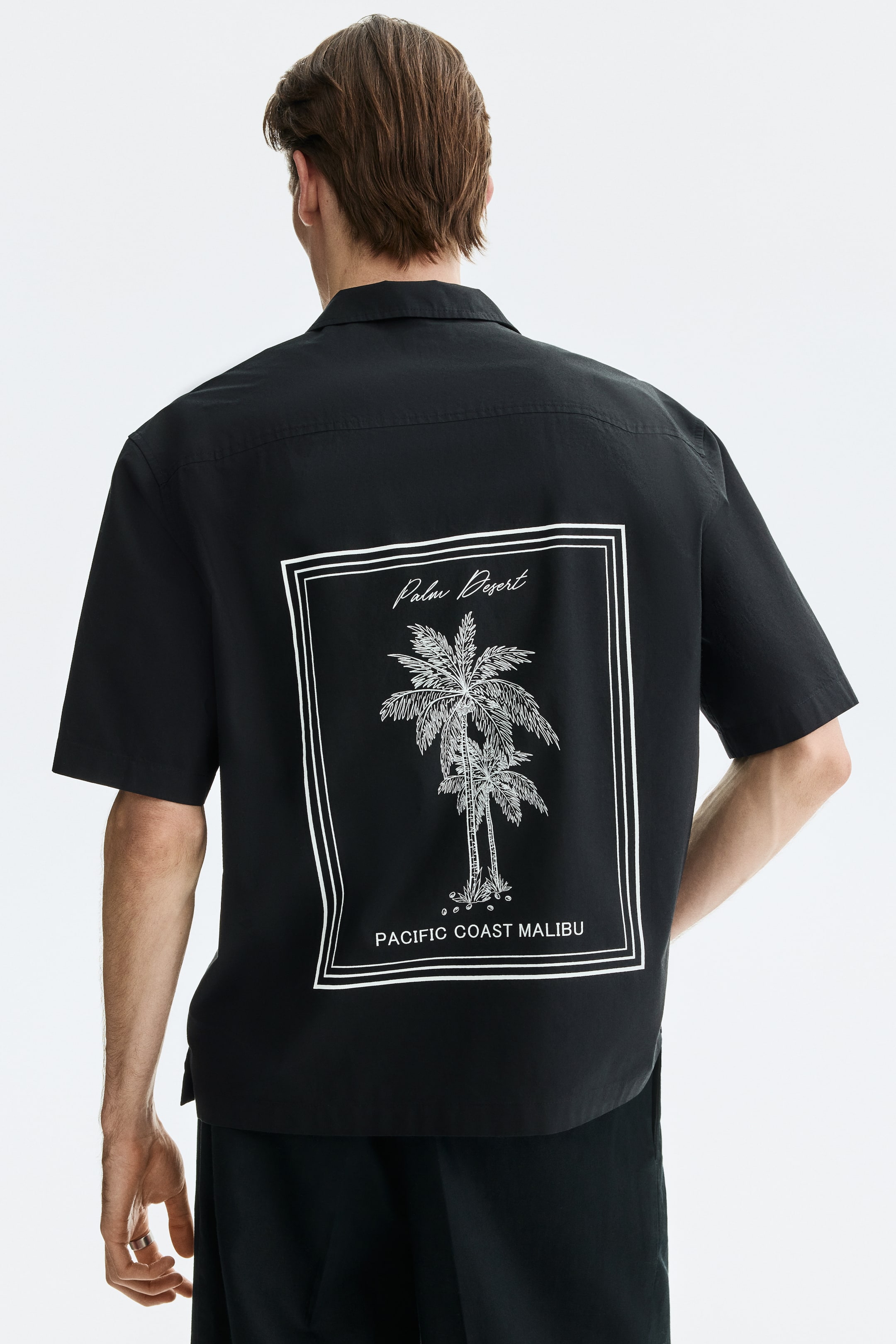 Größeres Bild anzeigen: Ein schwarzes kurzärmeliges Shirt wird von hinten betrachtet, mit einem weißen Grafikdruck auf der Rückseite. Der Druck zeigt zwei Palmen, die in einem doppelten rechteckigen Rahmen eingeschlossen sind, mit "Palm Desert" darüber und "PACIFIC COAST MALIBU" darunter.