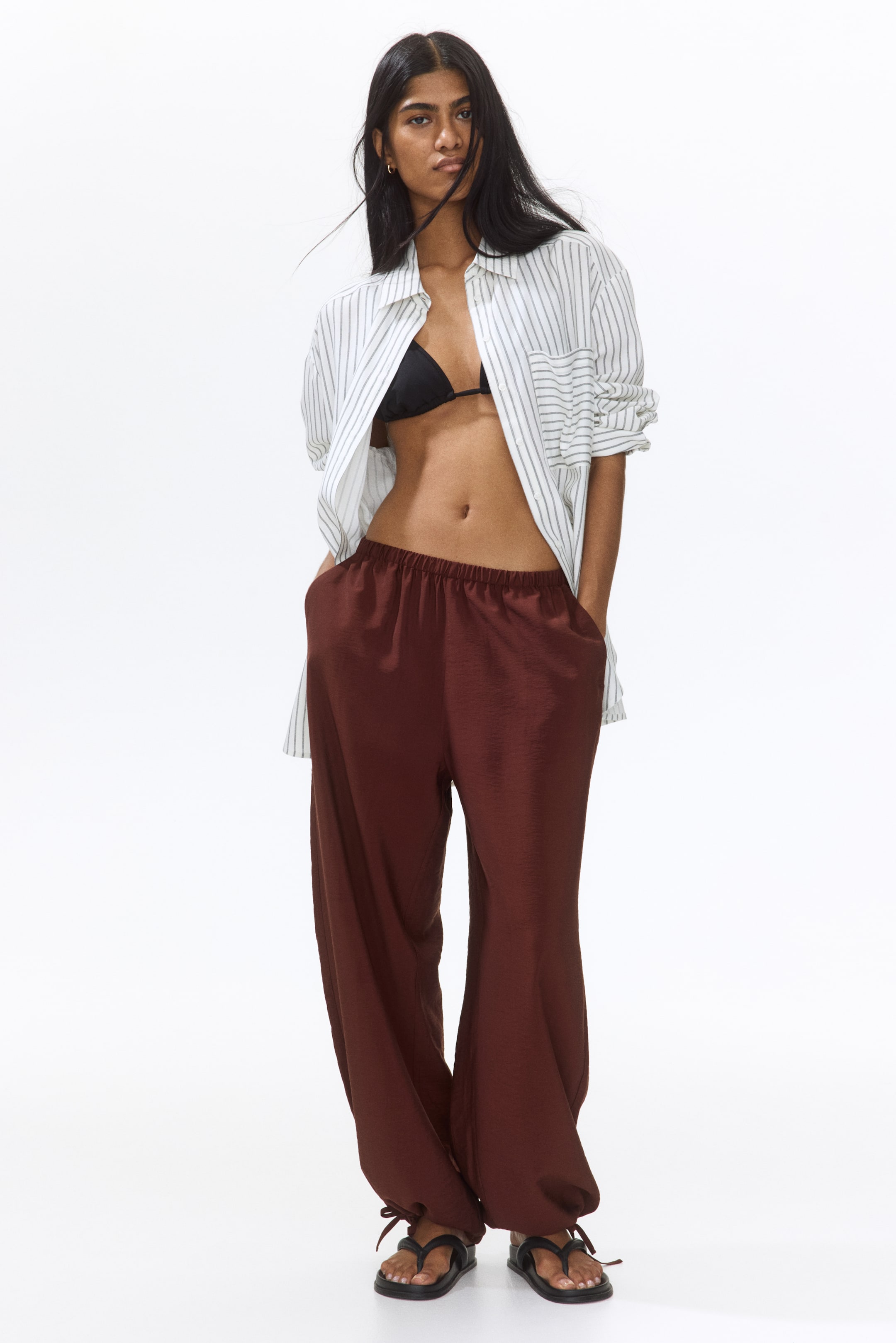 View larger image: Brown wide-leg pants are showcased from a slightly angled front perspective, highlighting an elasticated waistband and side pockets with hands comfortably placed in them. The flowing, textured fabric gathers at the ankles with tie details, crafting a relaxed silhouette.