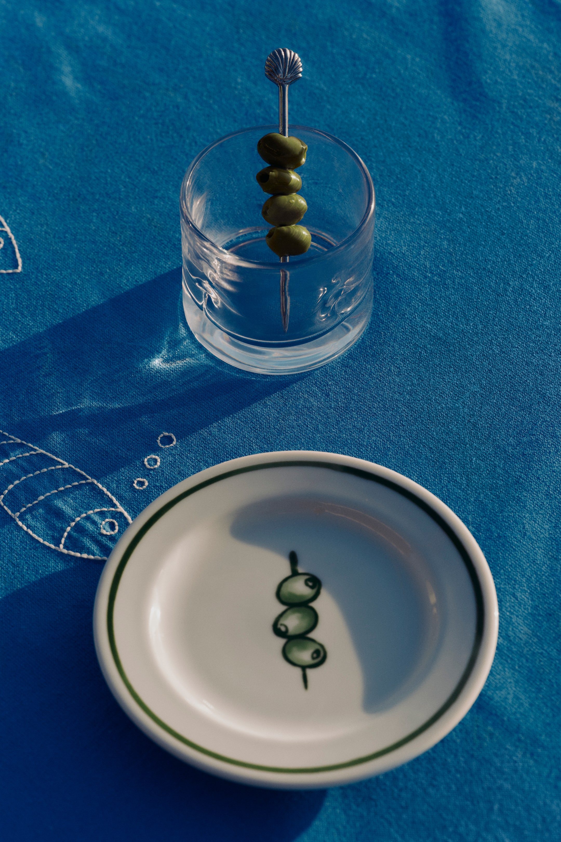 Ampliar la imagen: Un vaso de vidrio transparente sostiene un pincho de metal plateado, en el cual están ensartadas cuatro aceitunas verdes, con un diseño decorativo en forma de concha en la parte superior. Debajo, se encuentra un pequeño plato redondo de cerámica blanca, que presenta un borde verde oscuro y un diseño pintado de tres aceitunas verdes en un tallo.