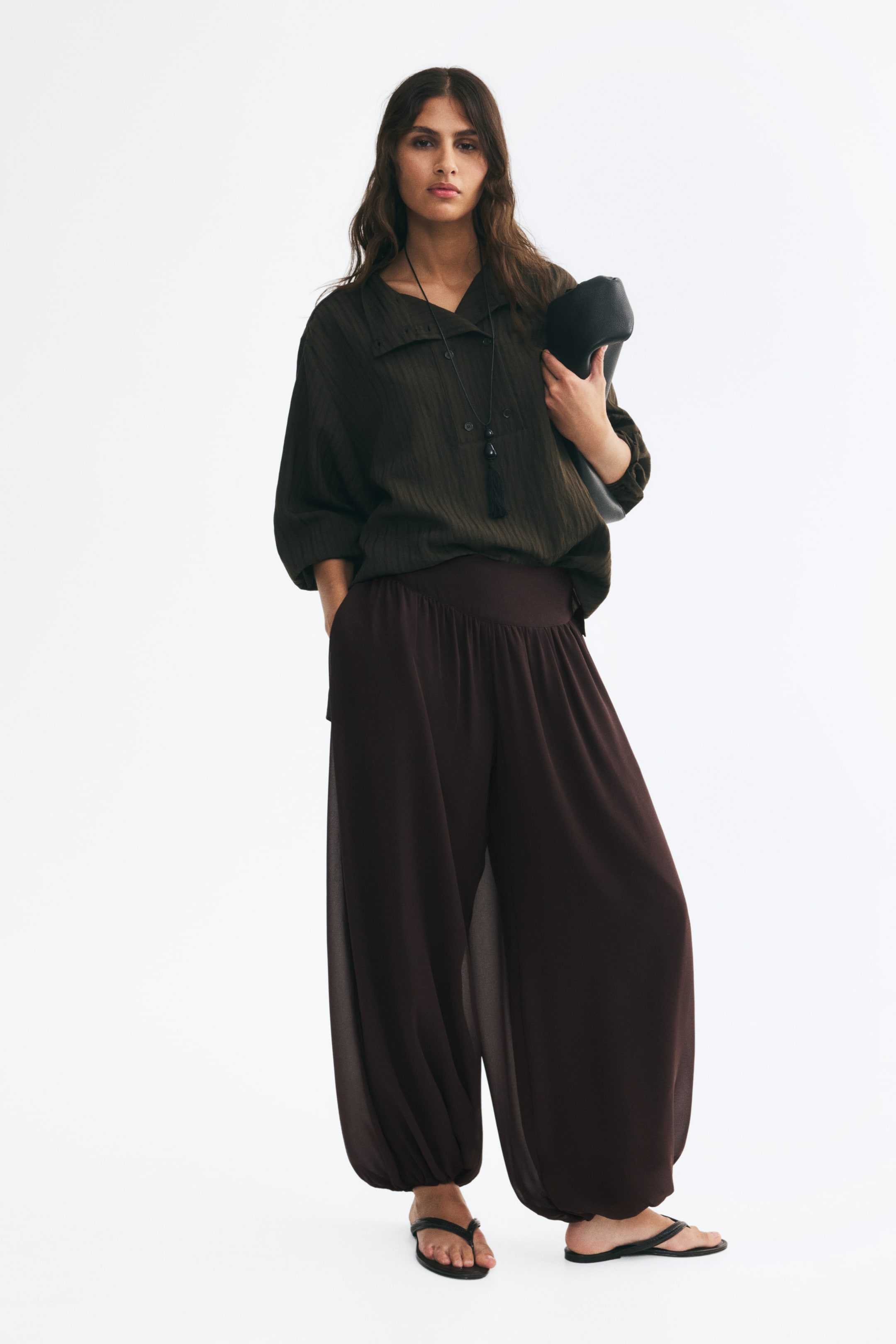 View larger image: Dark brown, wide-leg harem trousers with a gathered waistband and cuffs are worn by the model, who stands with one hand tucked into a pocket. The trousers are styled with a loose-fitting, dark green textured tunic and a black leather clutch tucked under the arm.