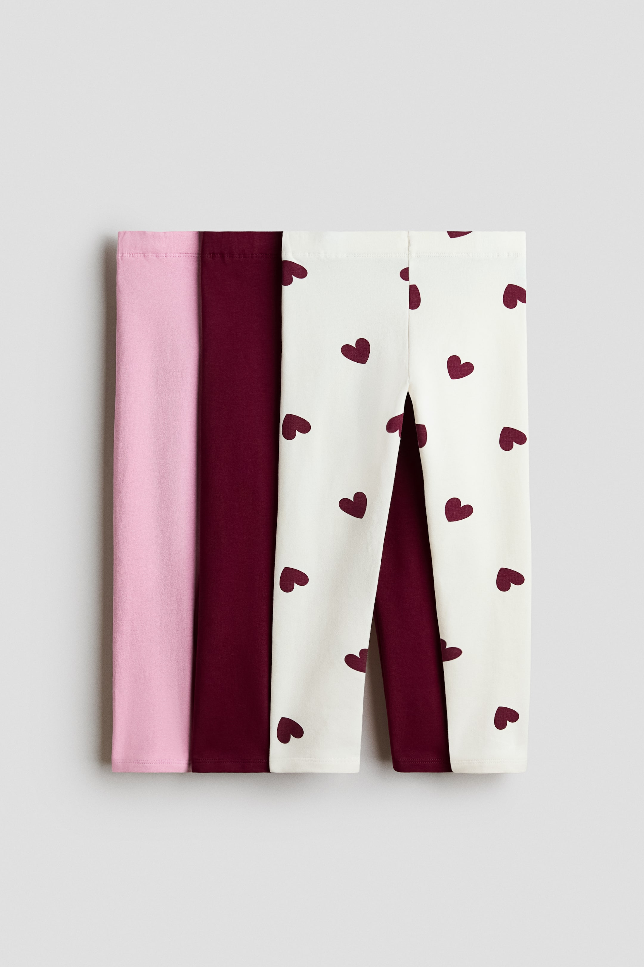 Voir une image plus grande: Trois paires de leggings sont disposées à plat et partiellement superposées, visibles d’un point de vue en plongée. Une paire est rose pâle, une est d’un pourpre rougeâtre foncé, et une est crème avec de petits cœurs pourpre rougeâtre foncé.