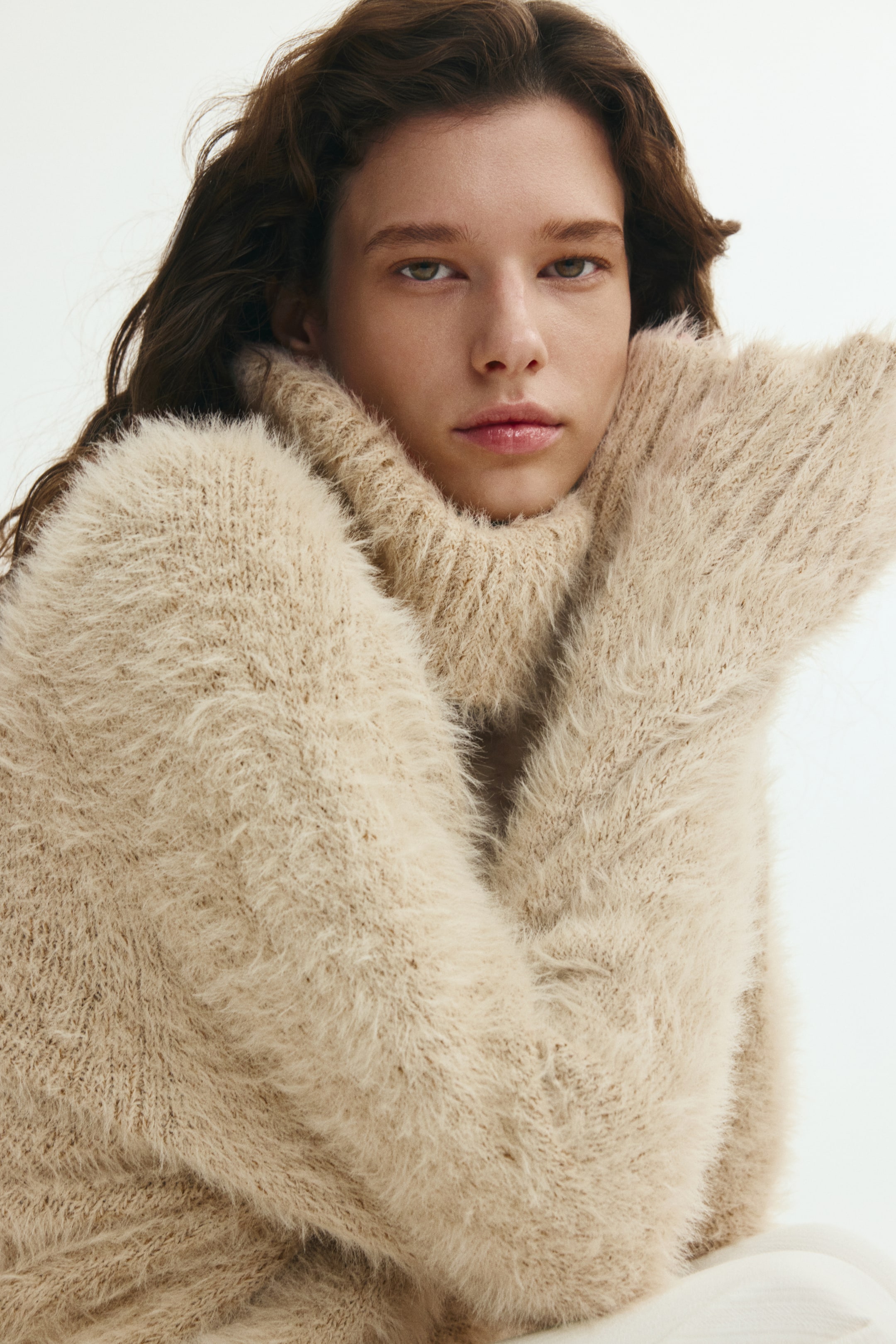 View larger image: The model is wearing a beige sweater in a soft, fluffy knit, resting their chin on their sleeve-covered hands. The sweater features a thick ribbed turtleneck and long textured sleeves.