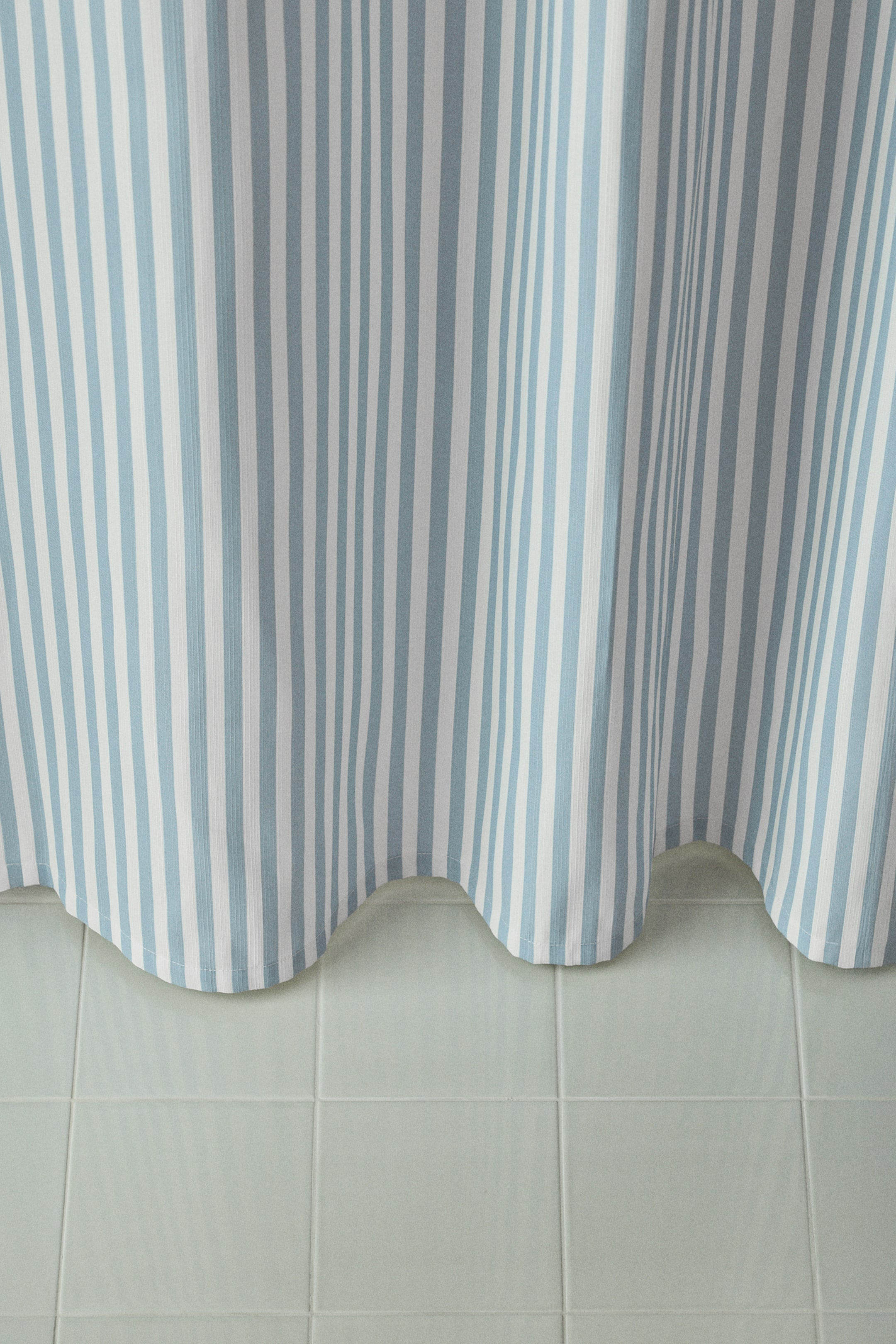View larger image: A vertically striped curtain is hanging, featuring alternating light blue and white stripes. The fabric is forming soft, vertical folds, creating a wavy, undulating hemline.